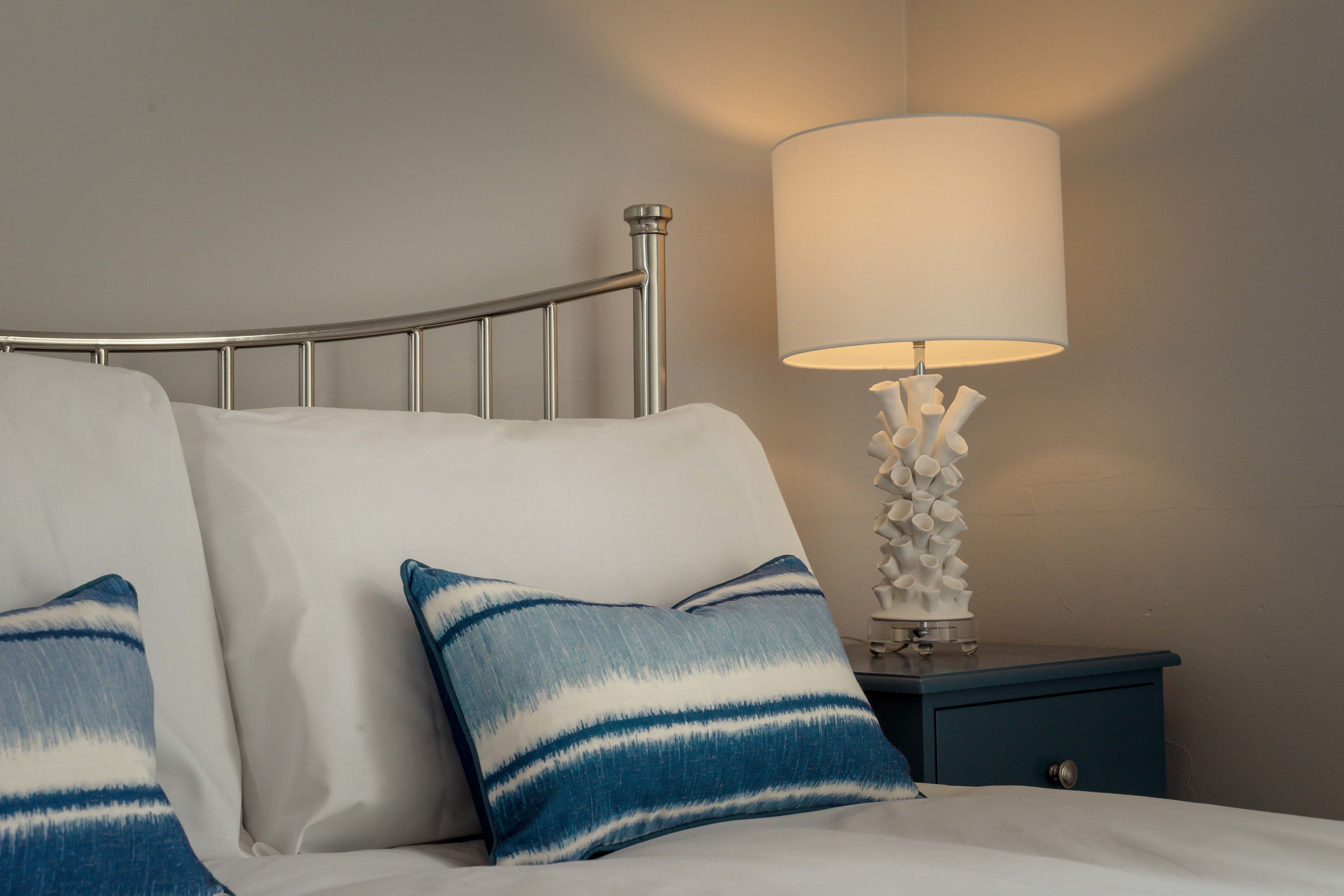 Ein ordentlich bezogenes Bett mit blau-weiß gestreiften Kissen, einem Metall-Kopfschild und einer weißen Lampe mit ungewöhnlichem Fuß auf einem blauen Nachttisch.