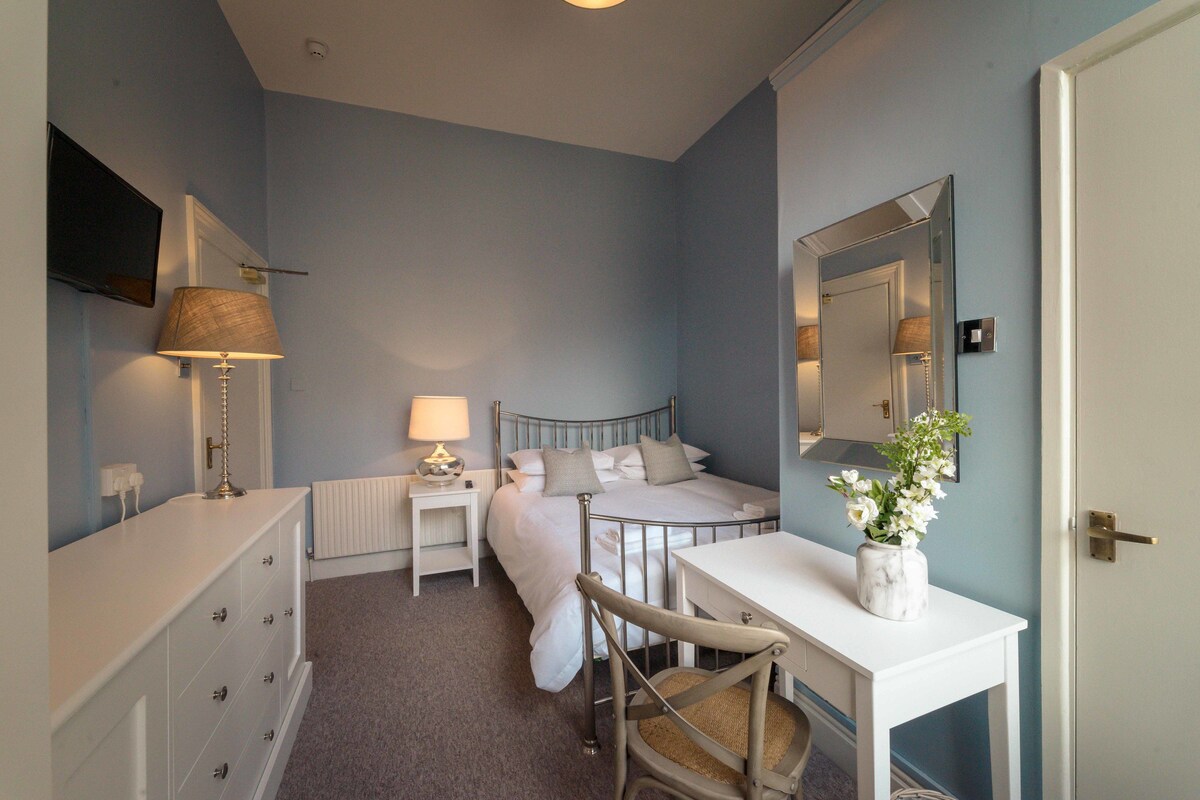 A bedroom with a bed, bedside table, lamp, dresser, and a mirror on the wall.