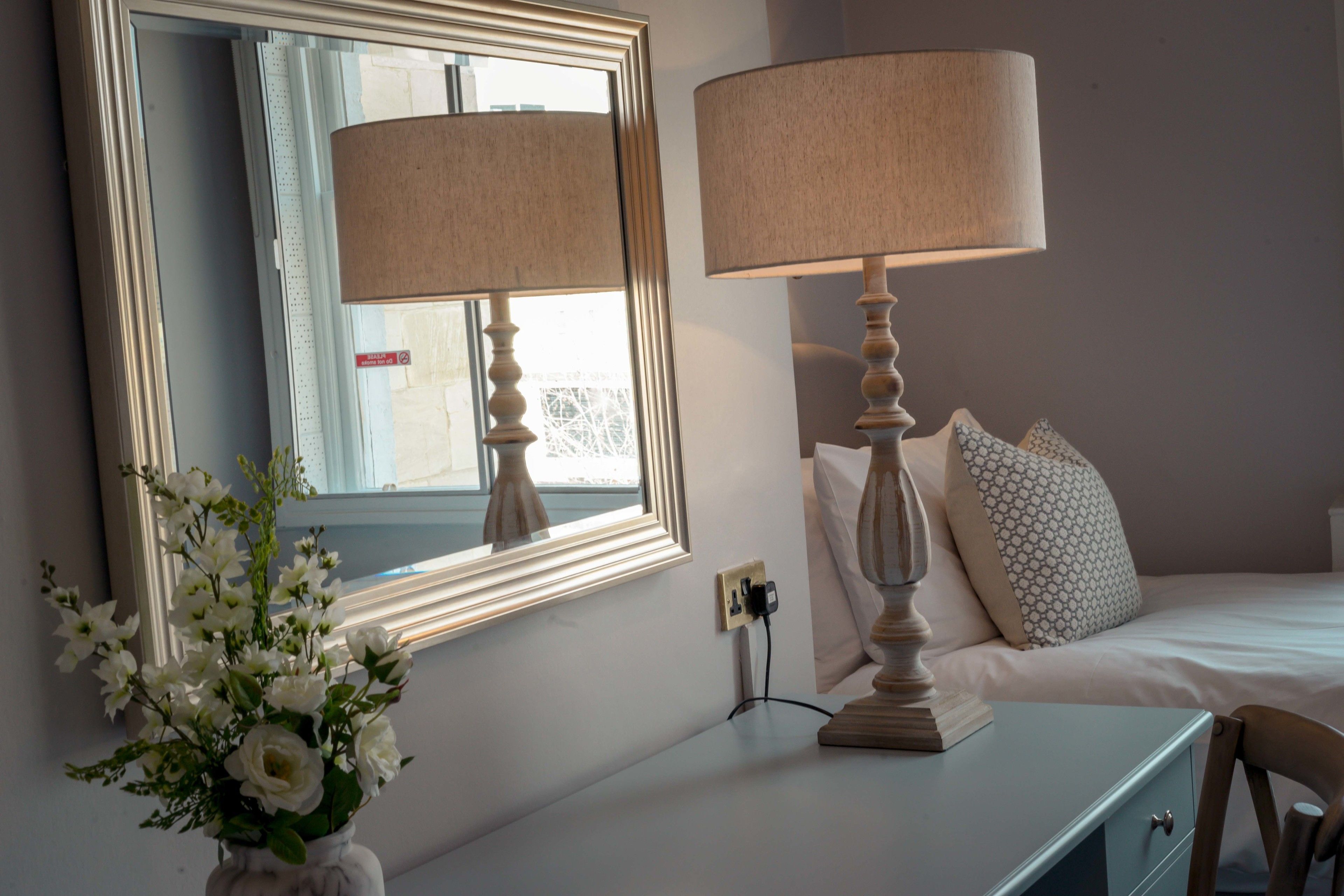 A bedroom with a bed, a bedside table with a lamp, a mirror, and a vase with flowers.