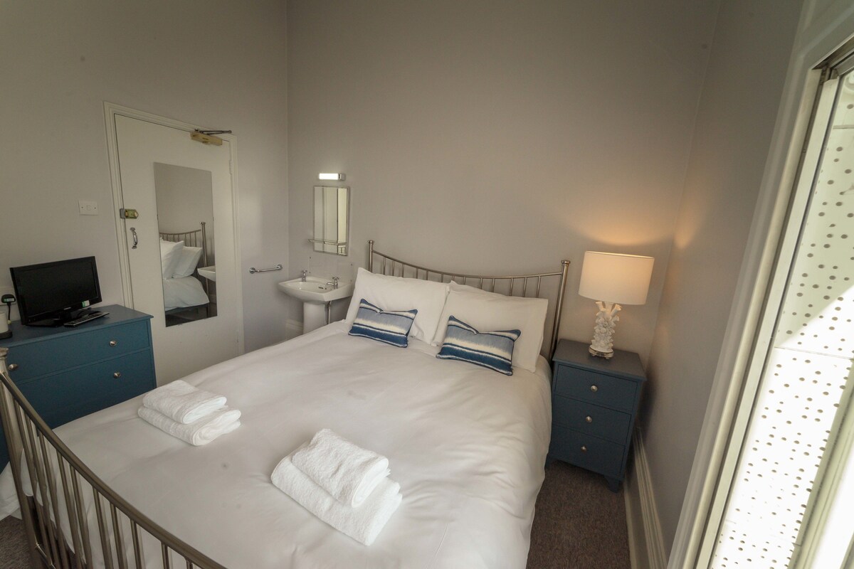 A neatly made bed with white linens and two pillows, a bedside table with a lamp, a small bedside table with a television, a mirror, and a door.