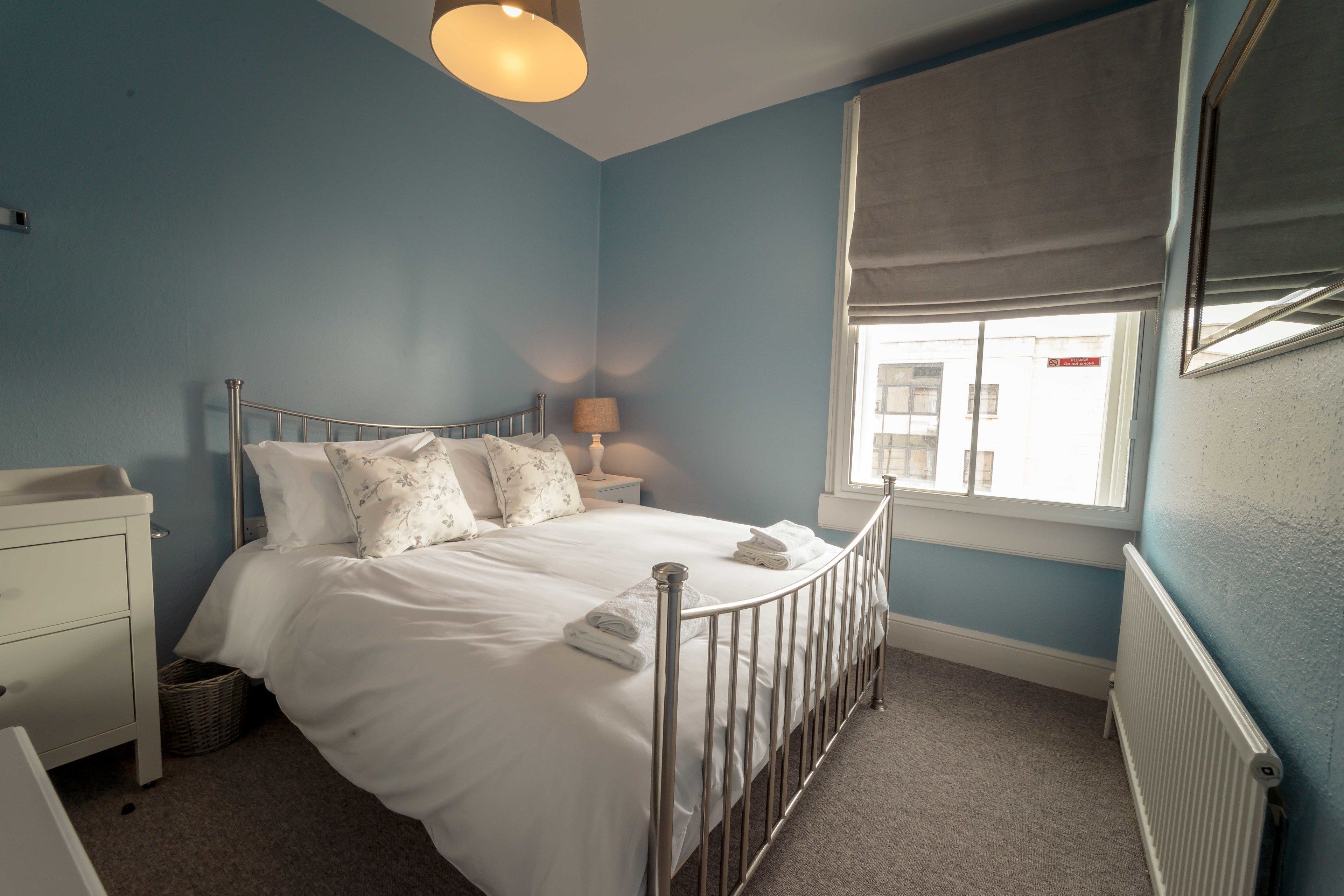 A bedroom with a metal bed frame, white bedding, a bedside table with a lamp, a window with a blind, and a radiator.