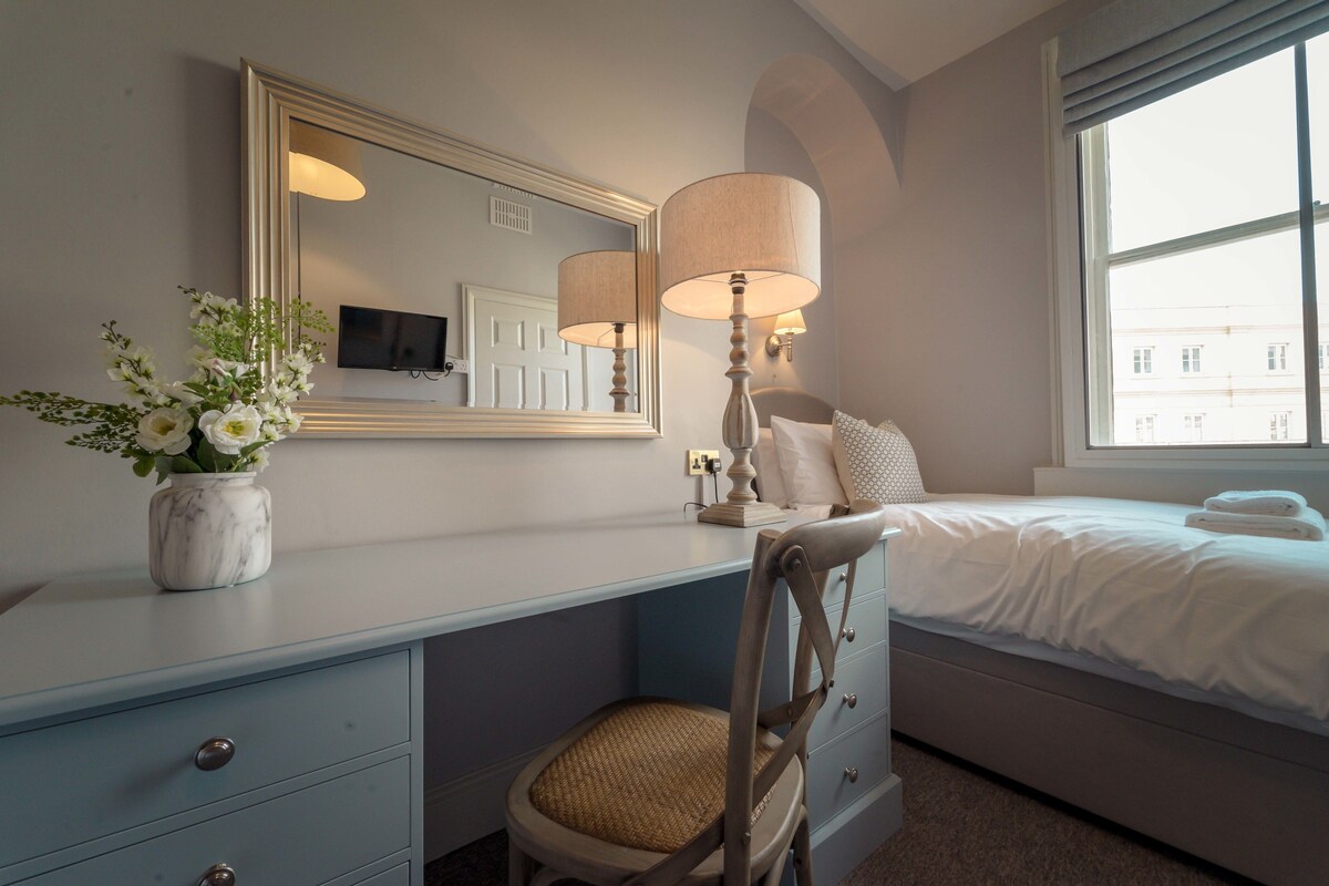 A hotel room with a bed, a desk with a chair, a mirror, a lamp, and a vase with flowers.