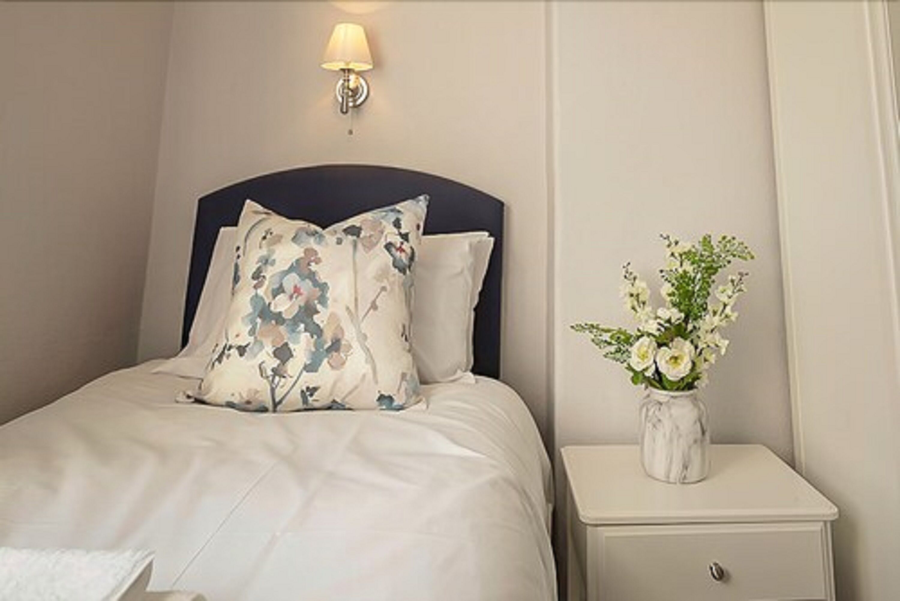 A neatly made bed with floral pillows, a bedside table with a vase of flowers, and a wall-mounted lamp.