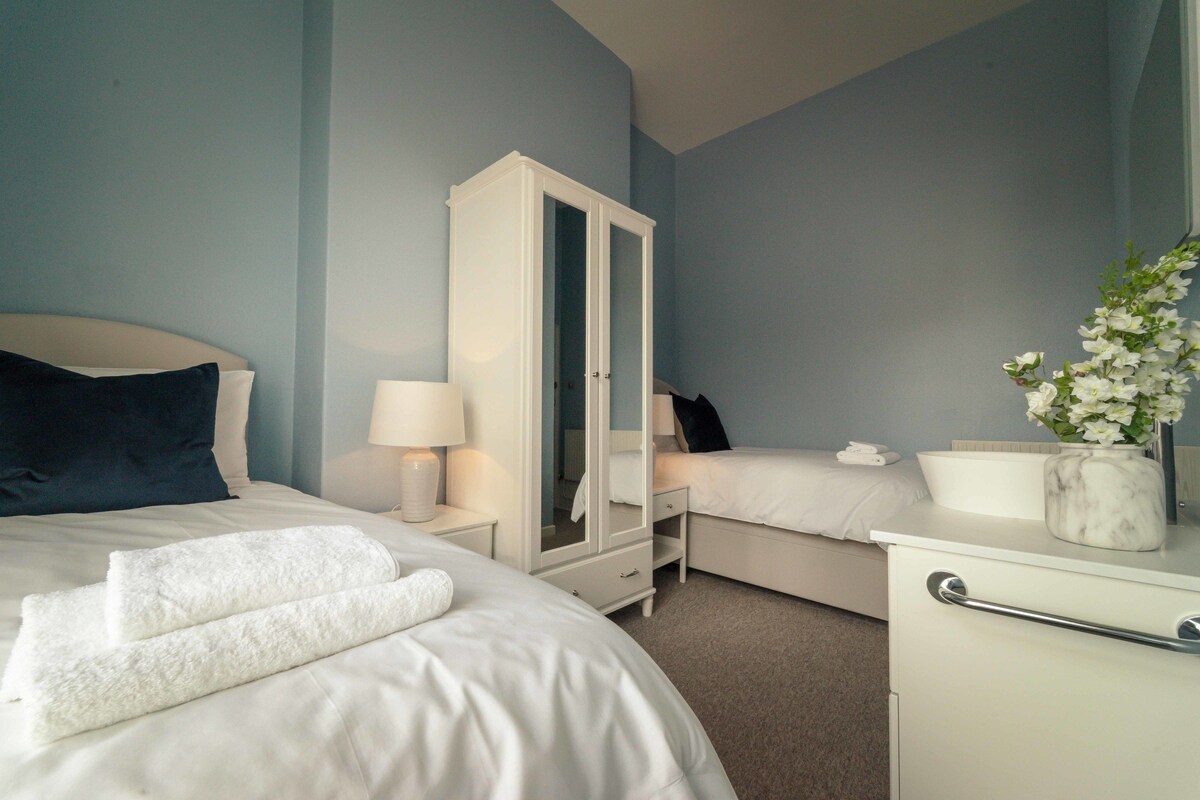 A hotel room with a bed, a wardrobe, a bedside table with a lamp, a small desk with a vase of flowers, and a nightstand with a phone.