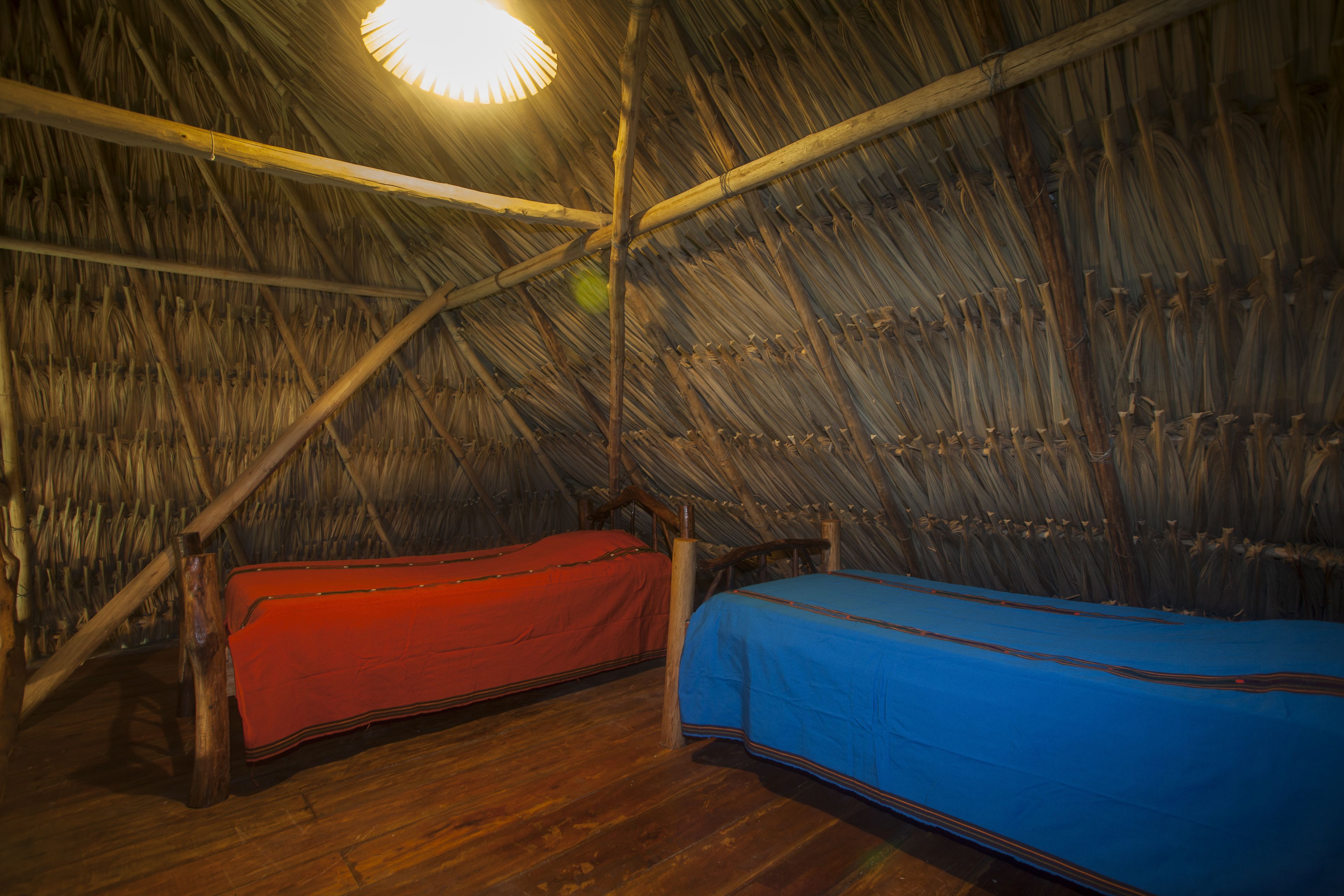 A room with a thatched roof, wooden beams, and two beds with red and blue bedding.