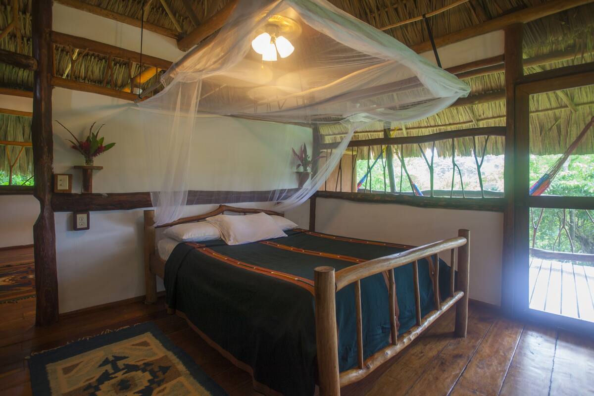 A wooden bed with a mosquito net, a potted plant, and a view of the outdoors.