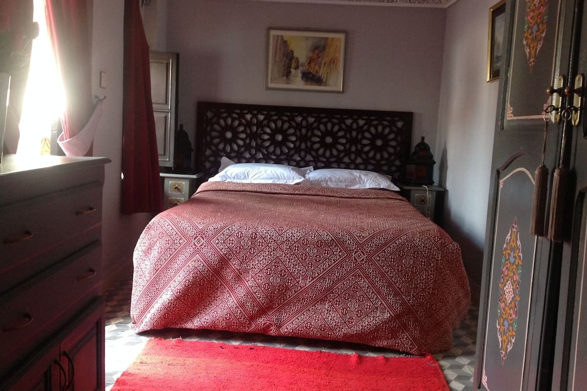 A bedroom with a patterned bedspread, a wooden headboard, a nightstand with a lamp, and a red curtain.