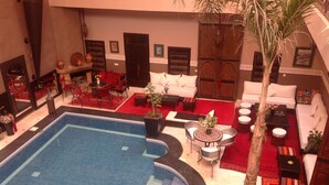 Un hall de hotel con piscina, sofás rojos y blancos, una palmera y una alfombra roja.