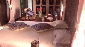 Habitación de hotel con una cama grande, dos sillas, una mesita con un jarrón de flores y vistas a un edificio tradicional con puertas decorativas.