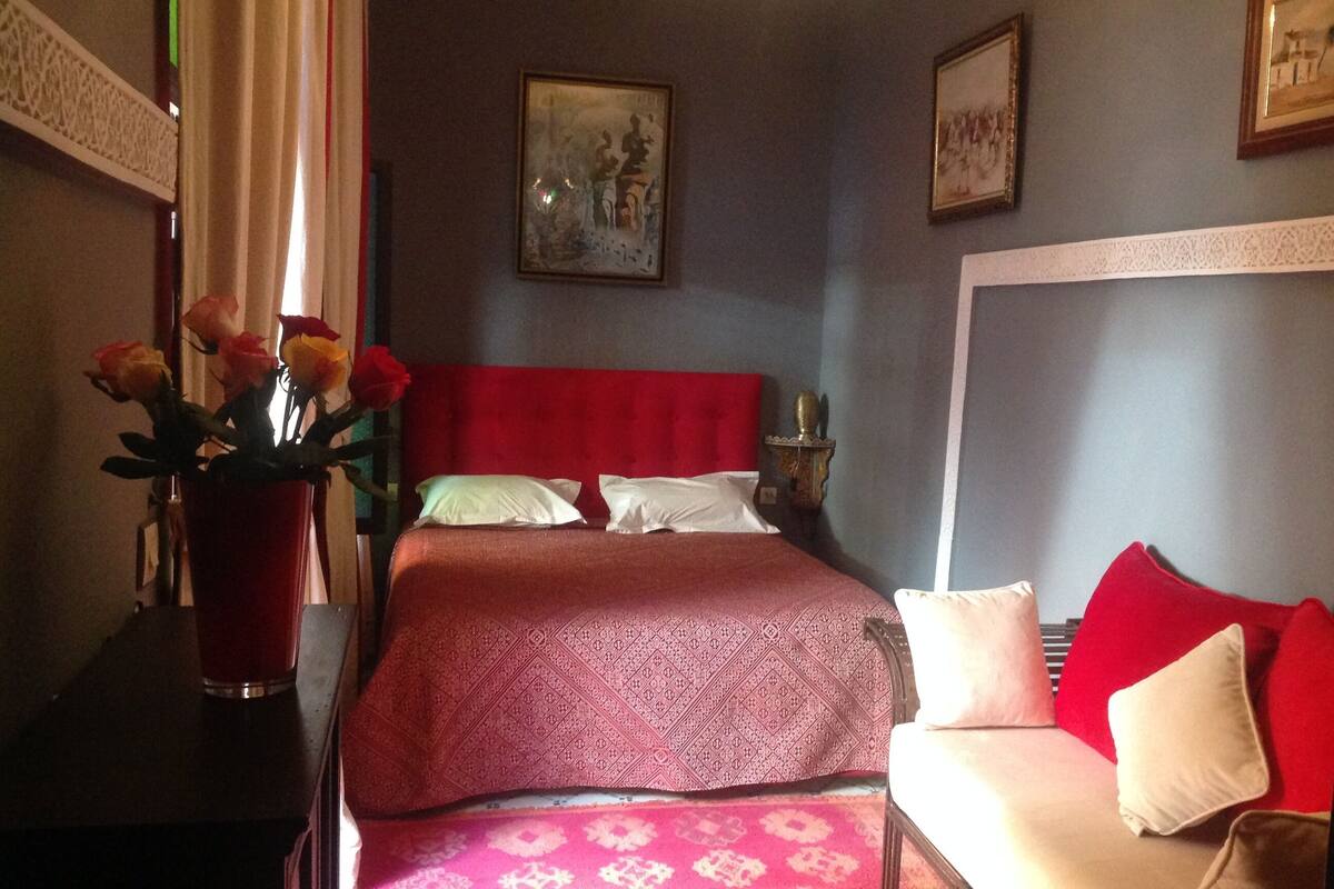 A bedroom with a red headboard, a bed with patterned bedding, a red sofa, a vase with flowers, and a framed picture on the wall.