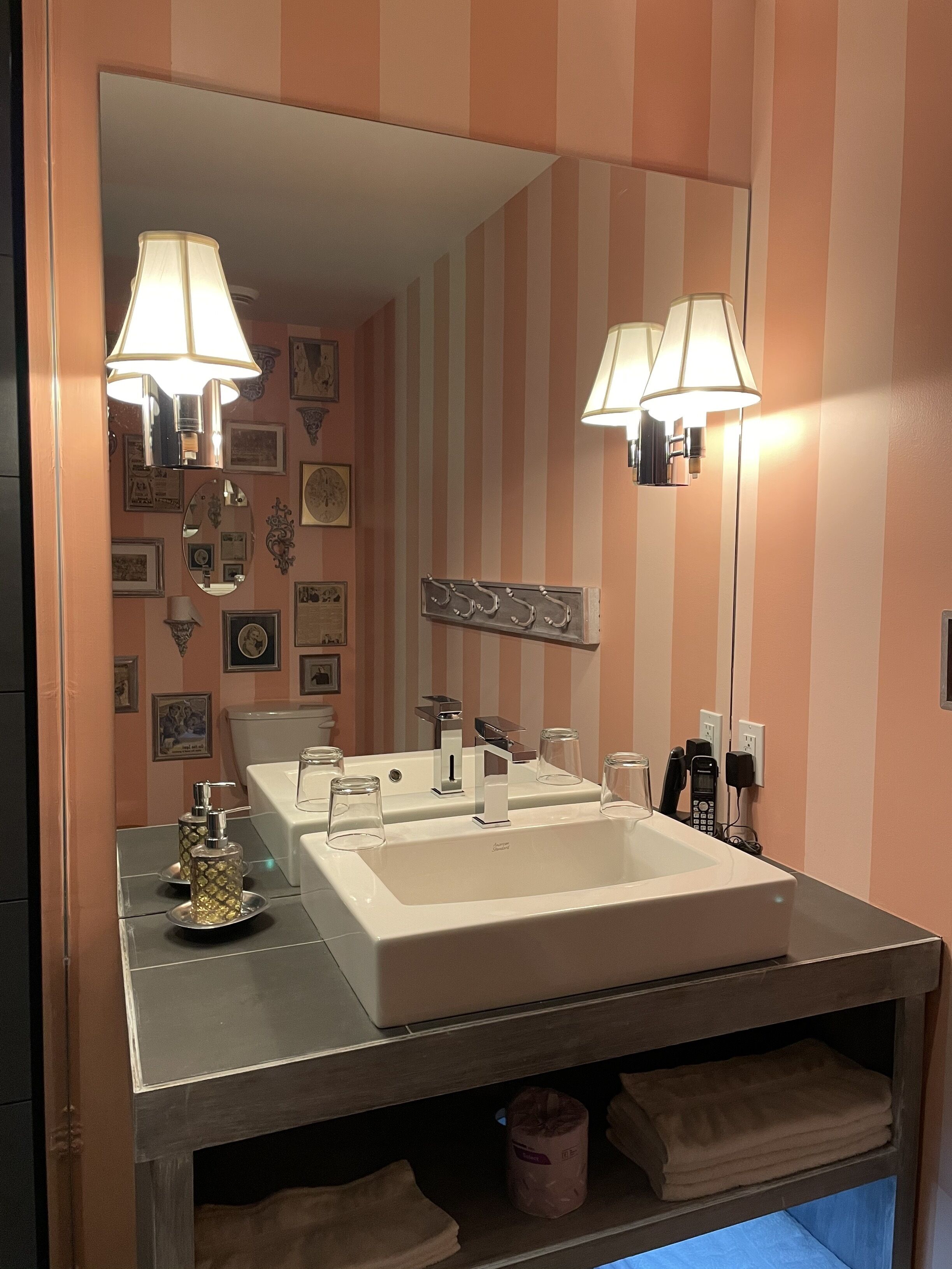 A bathroom with a large mirror, two wall-mounted lamps, a sink with a vanity, and a towel rack.