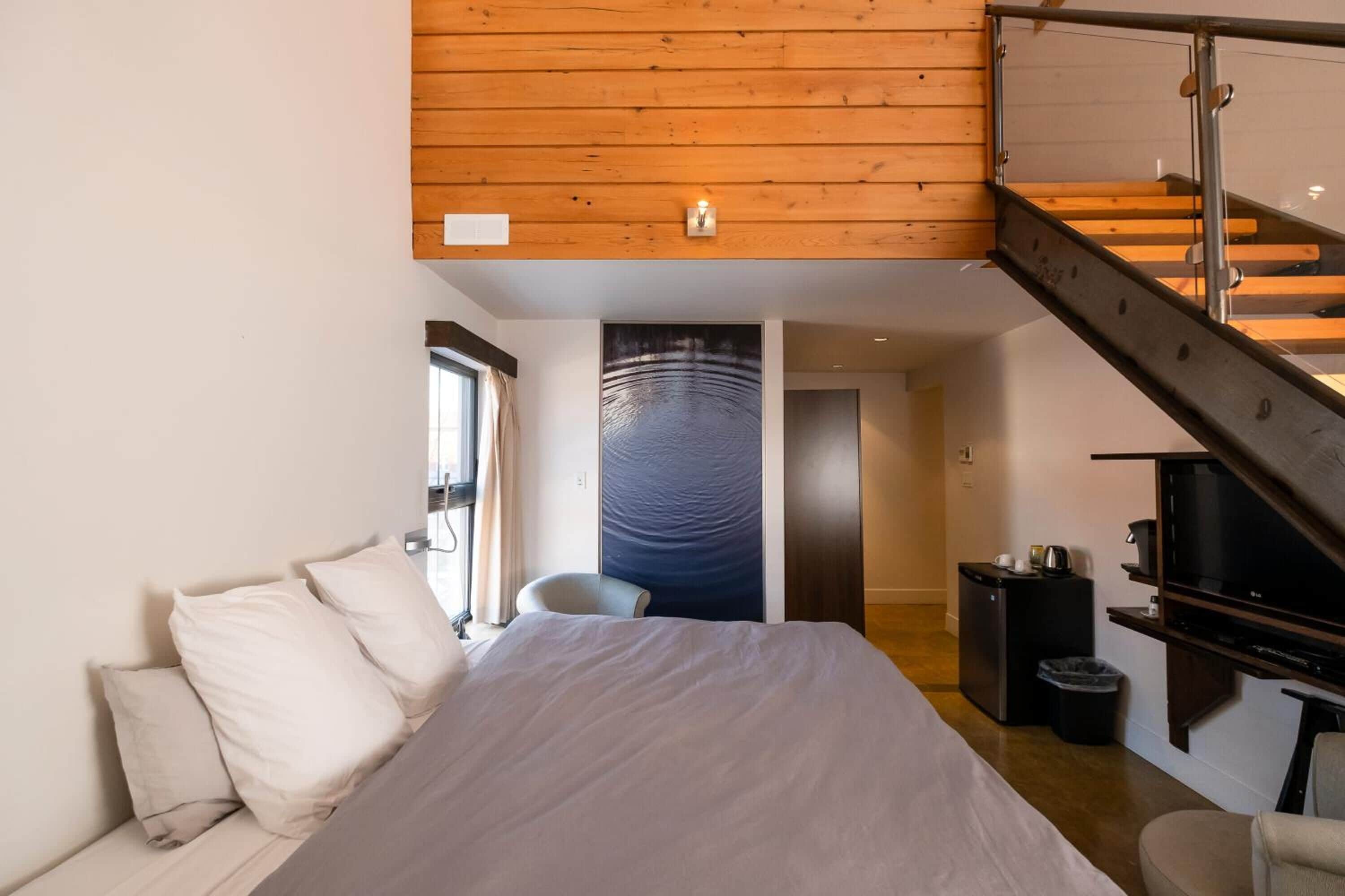 Una habitación de hotel moderna con una cama grande, techo de madera, una escalera, un escritorio pequeño y un televisor.