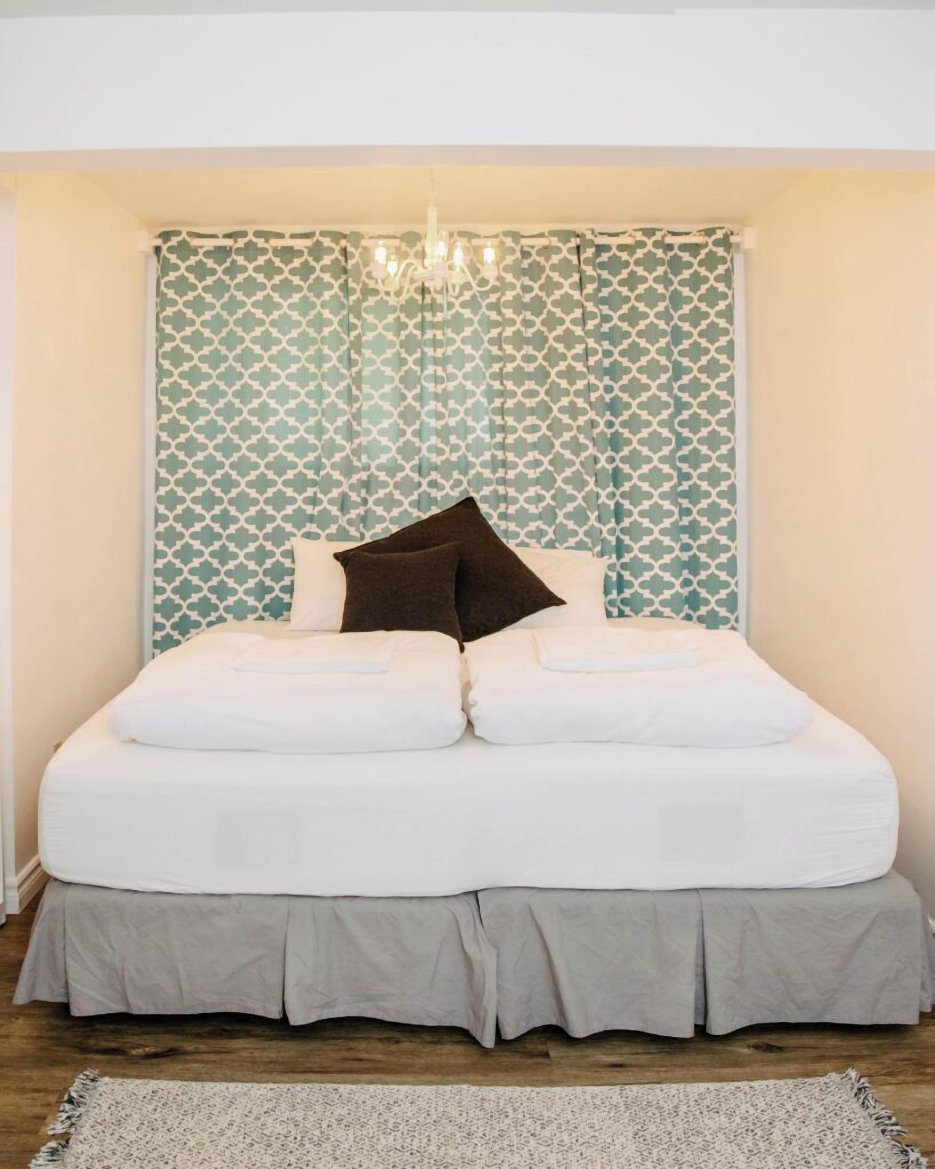 Una cama con ropa de cama blanca y falda gris, una cortina estampada y un candelabro.