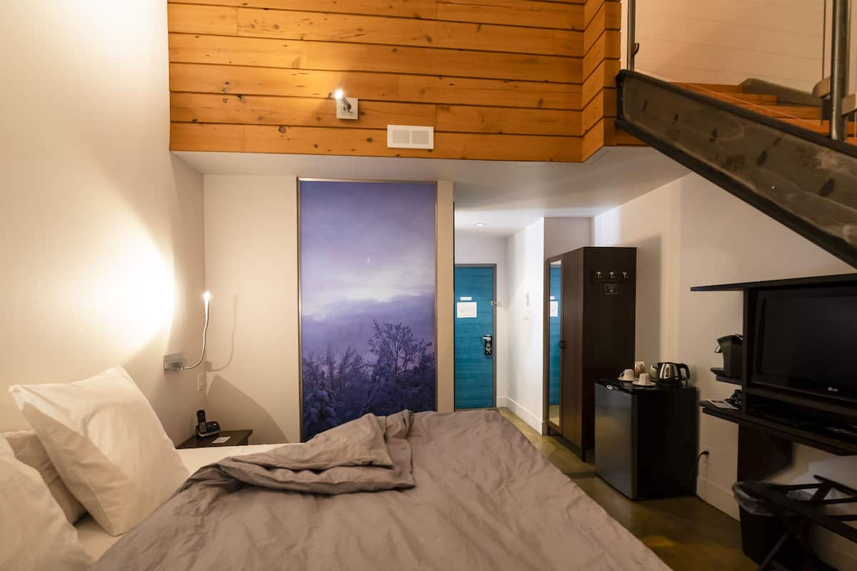 A bedroom with a bed, a nightstand, a wall-mounted lamp, a painting, a door, a small kitchenette, and a TV.