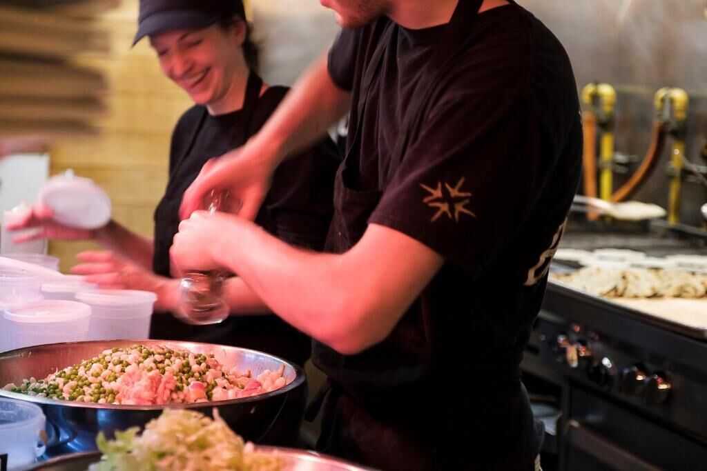 A kitchen area with chefs preparing food, a large metal bowl of chopped ingredients, and a stove in the background.