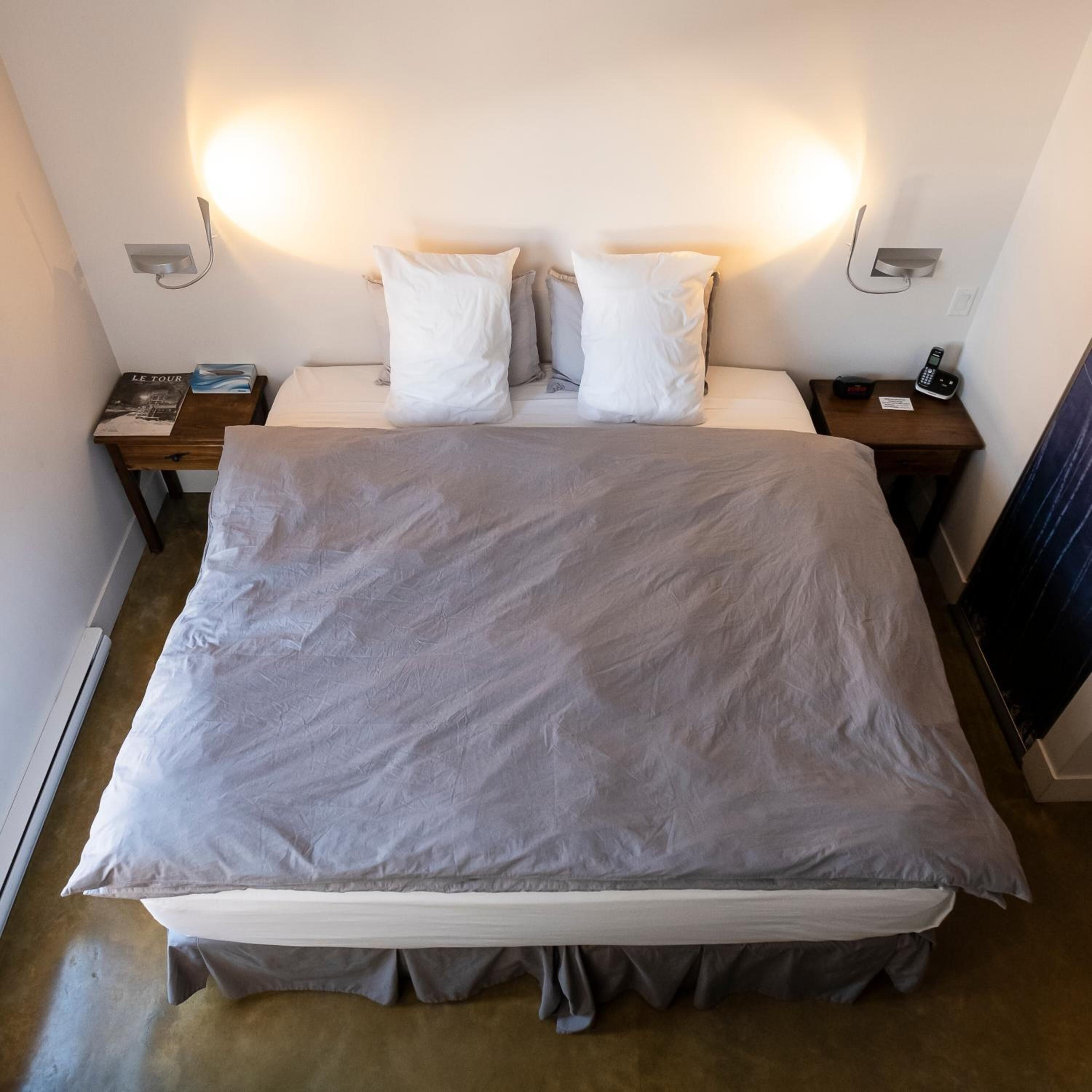 A neatly made bed with a grey bedspread and pillows, two bedside tables with lamps, and a telephone on one of the tables.