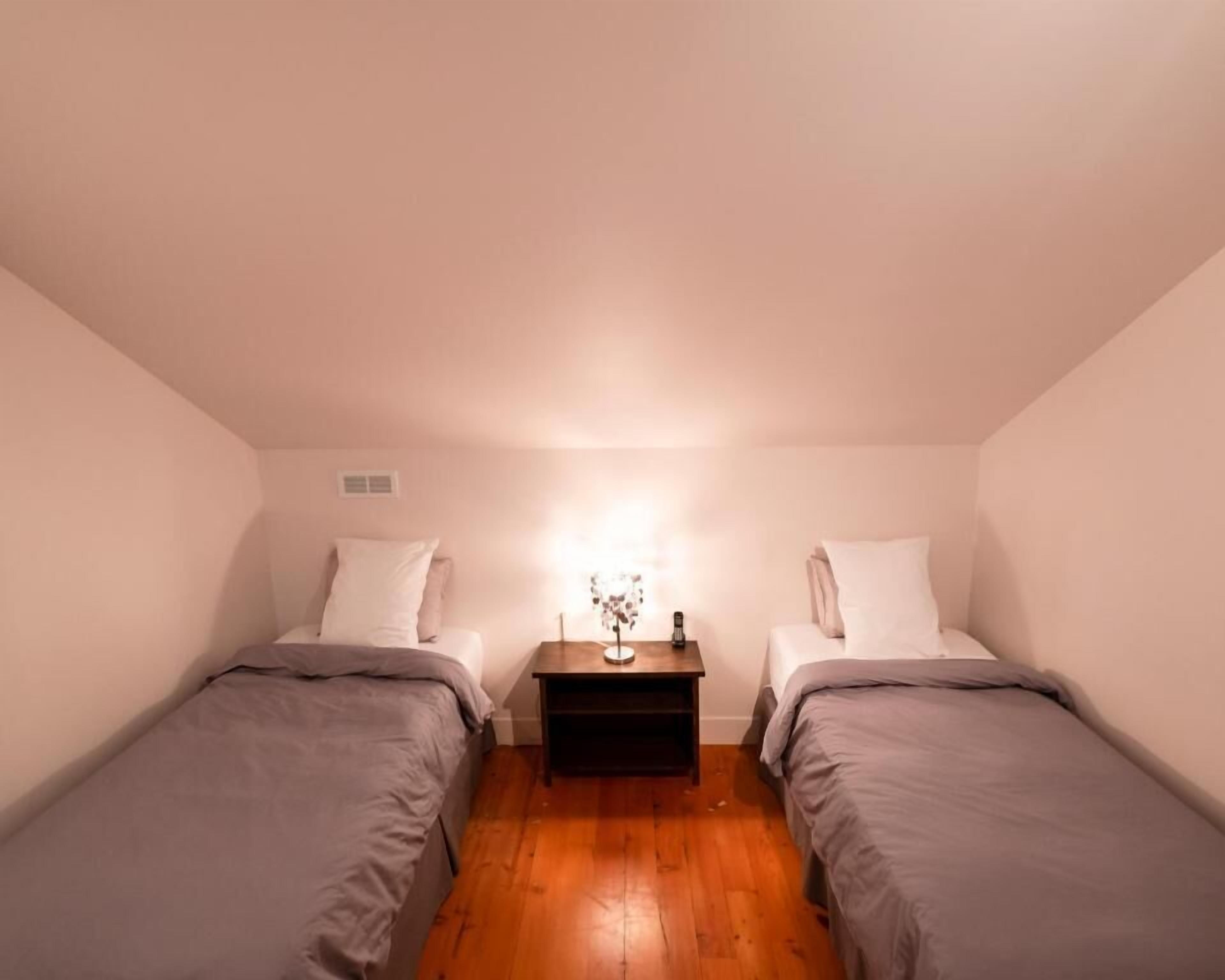 A small, dimly lit room with two single beds, a nightstand with a lamp, and a wooden floor.