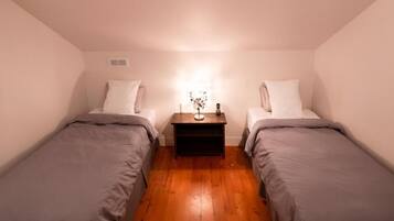 A small, dimly lit room with two single beds, a nightstand with a lamp, and a wooden floor.