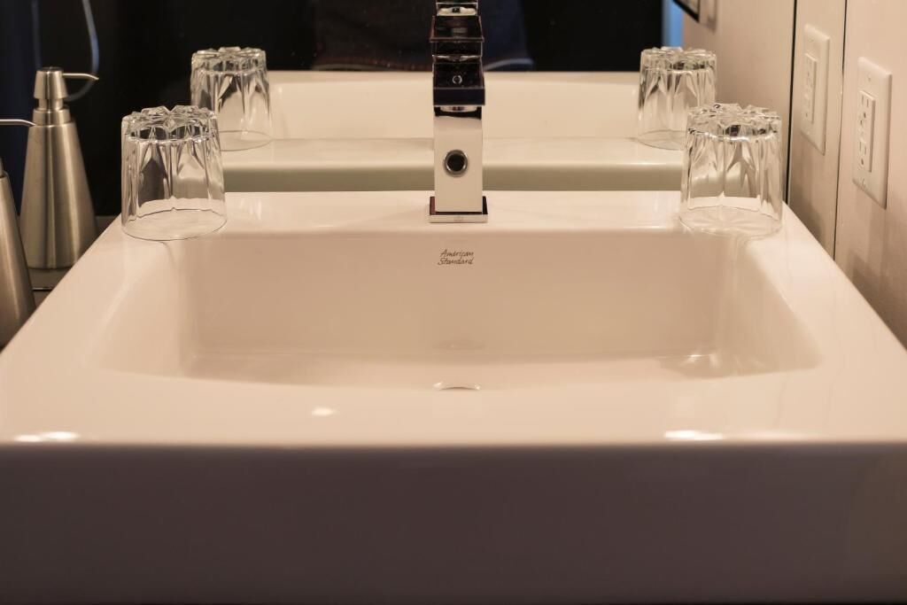 Un lavamanos blanco con grifo cromado, varios vasos de vidrio transparente y una botella de jabón para manos.