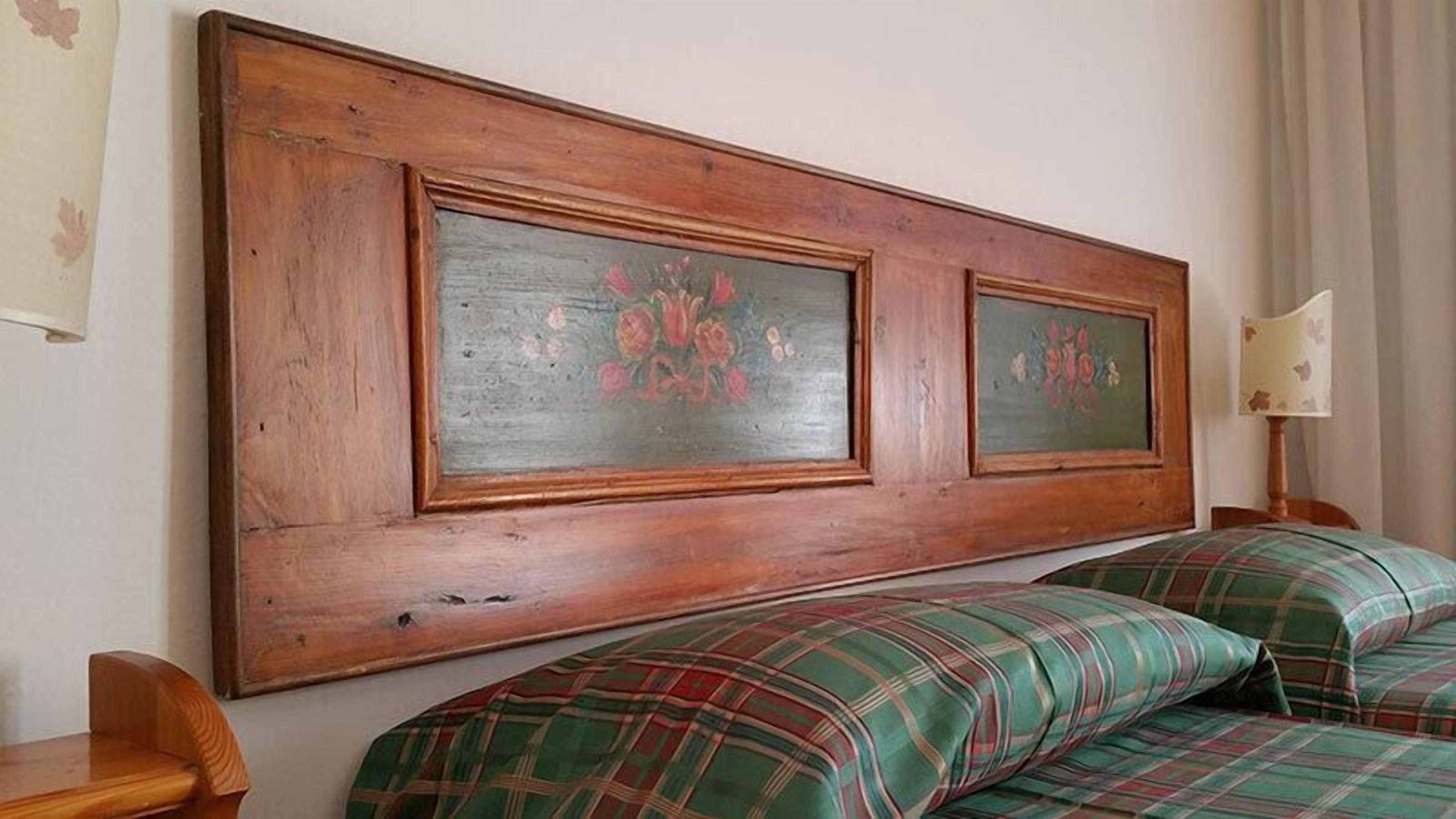 Una habitación con cabecera de madera, dos cuadros enmarcados con motivos florales, ropa de cama a cuadros, una mesita de noche de madera y una lámpara.