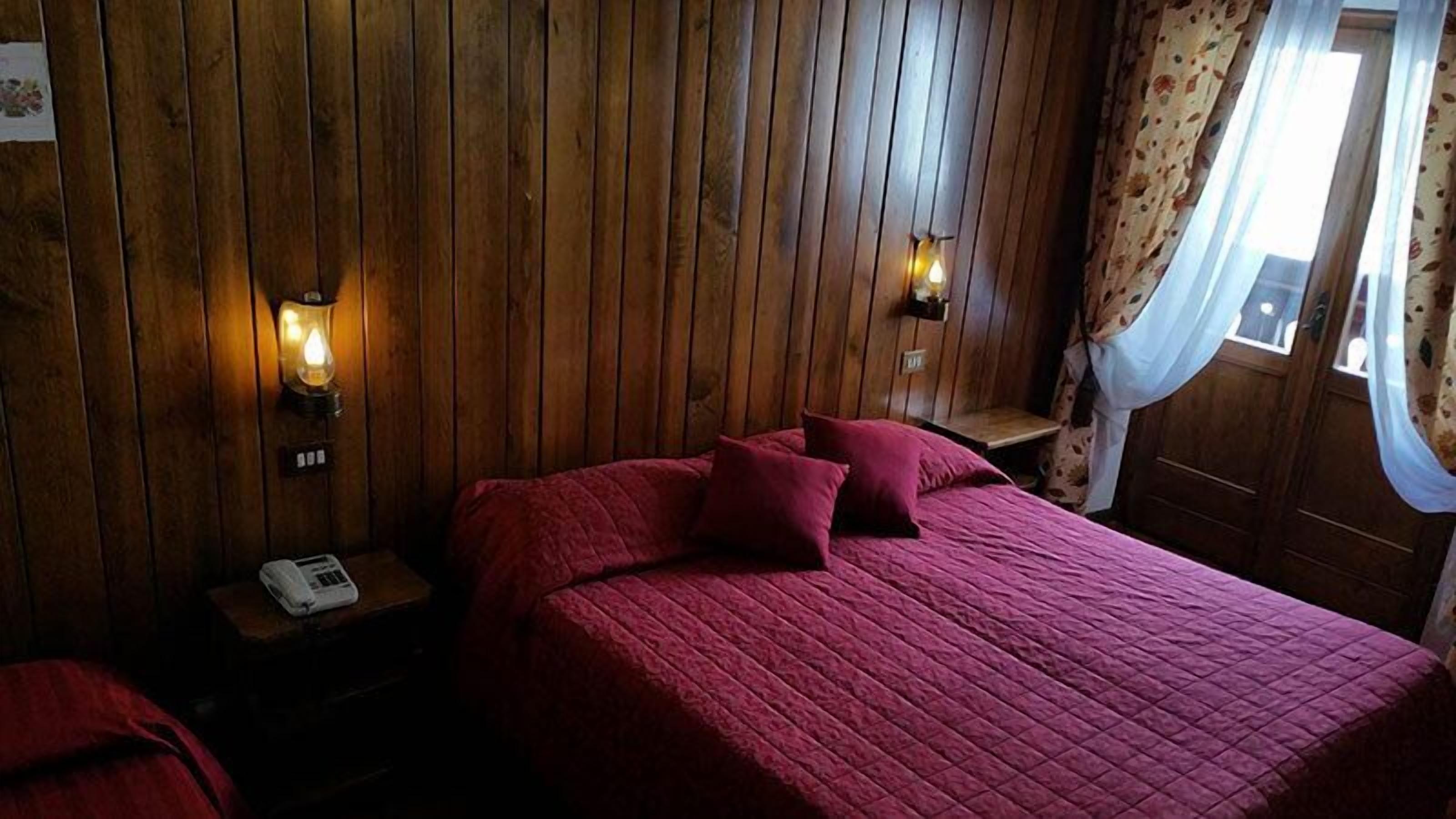 Una habitación con pared de paneles de madera, una cama con ropa de cama roja, dos lámparas fijadas a la pared y un teléfono en una mesita de noche.