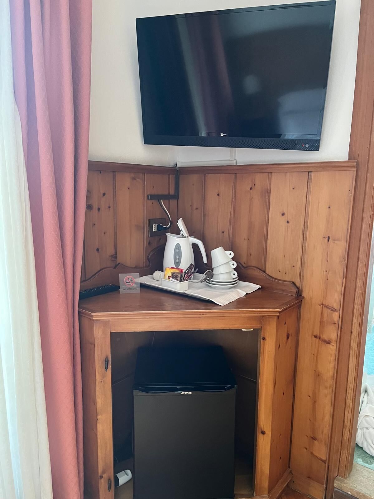 Ein Eckzimmer mit an der Wand montiertem Fernseher, einem kleinen Holztisch mit Wasserkocher, Tassen und einem Mülleimer.