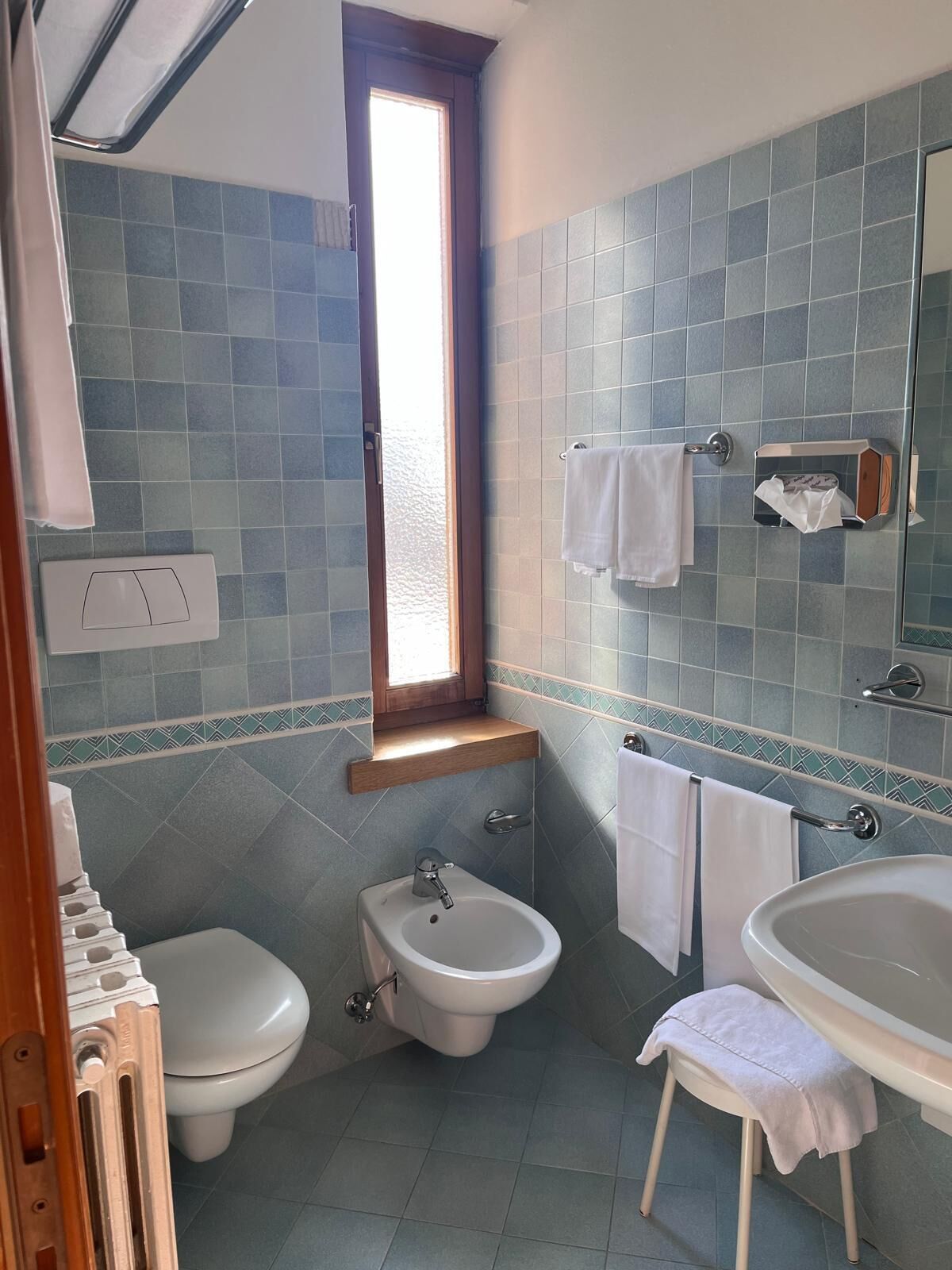 A bathroom with a toilet, bidet, and bathtub, tiled in blue and white, with a window and towel rack.