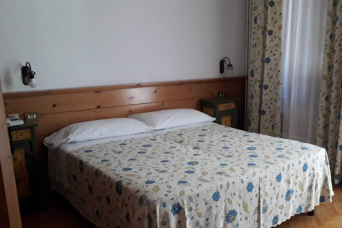 Un dormitori amb un capçal de llit de fusta, un llençol amb motius florals, dues taules de nit i una finestra amb cortines.