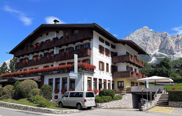 Un hotel de varios pisos con balcones decorados con flores rojas, un estacionamiento y montañas al fondo.