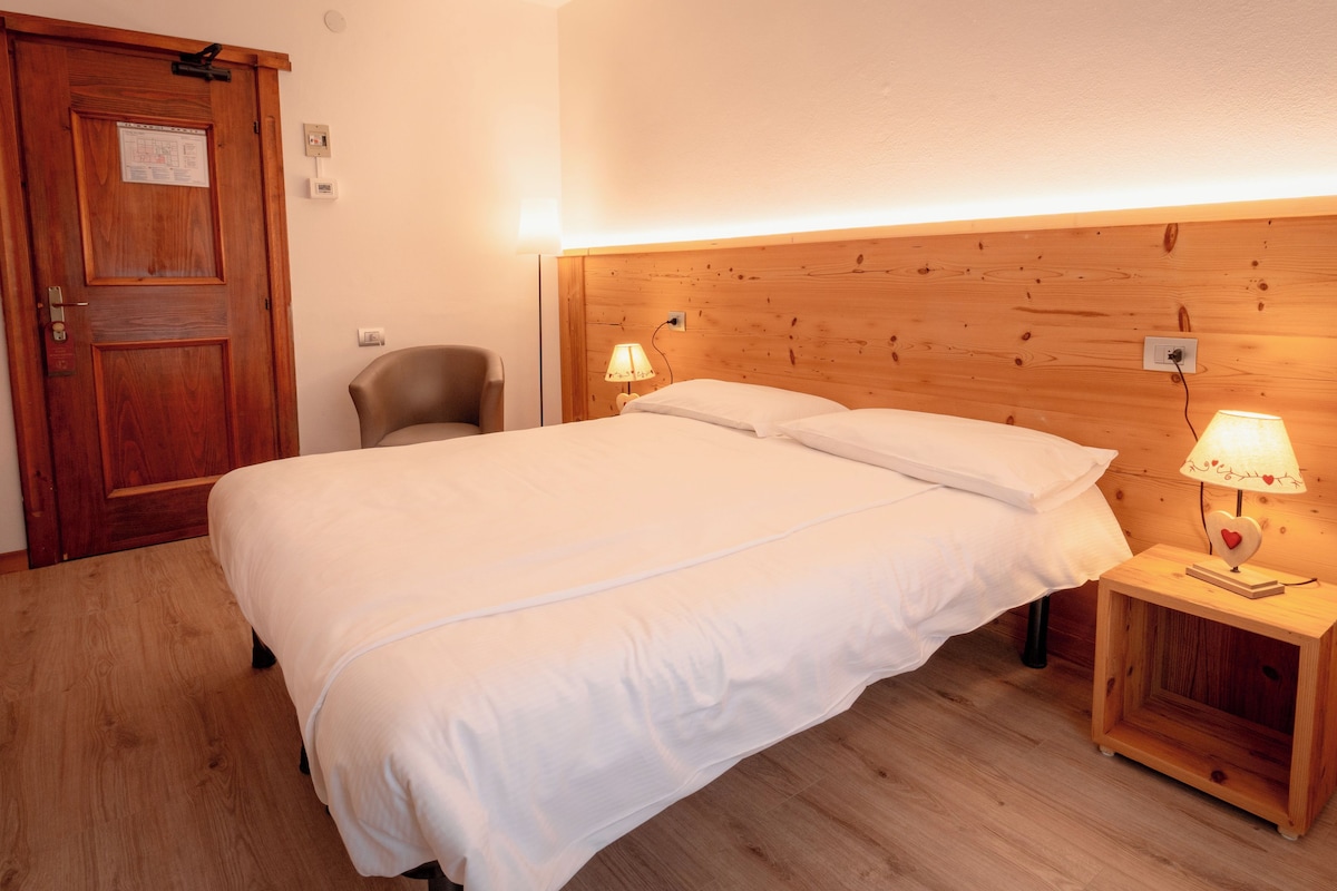 A hotel room with a wooden headboard, a bed with white linens, a chair, two bedside lamps, and a wooden nightstand with a small decorative item.