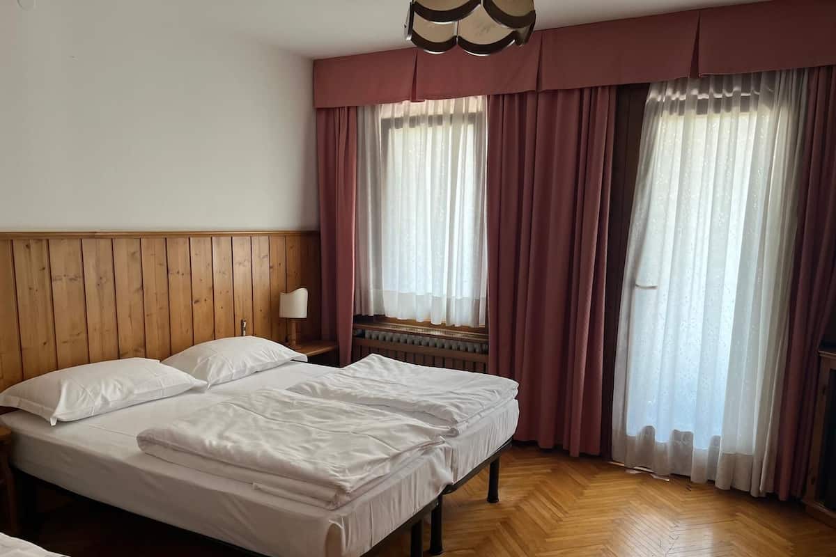 Habitació d'hotel amb dos llits, un panell de capçalera de fusta, una tauleta de nit amb una làmpada i grans finestres amb cortines.