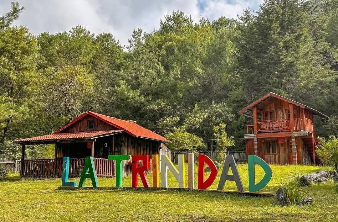 Eine Holzhütte mit rotem Dach und Balkon, umgeben von üppiger Vegetation. Ein Schild zeigt die Aufschrift "LA TRINIDAD".