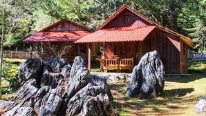 Una cabaña rústica con techo rojo, rodeada de grandes rocas y un bosque denso.