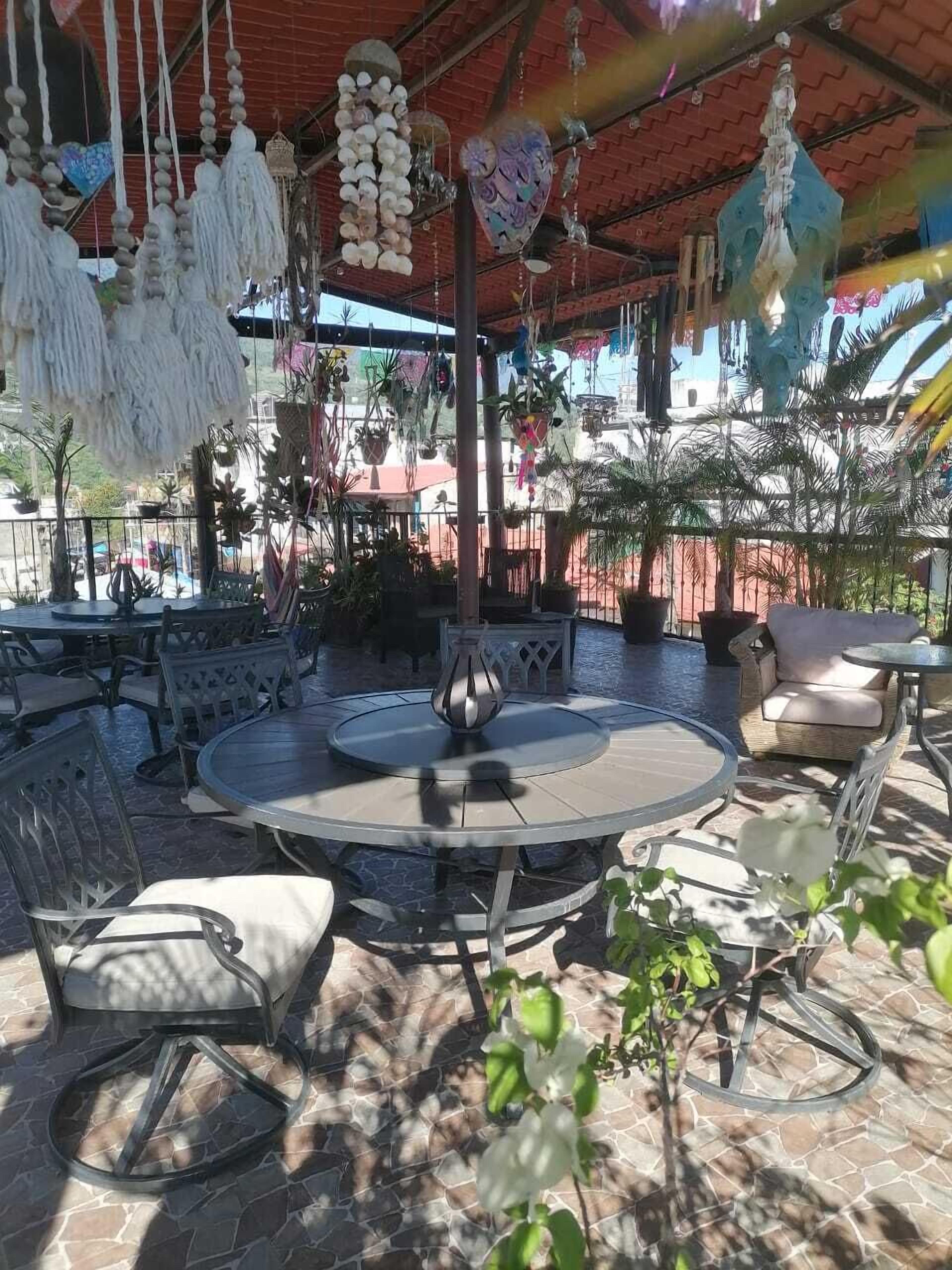 Un espacio al aire libre con mesas y sillas redondas, rodeado de plantas en macetas y adornos colgantes.