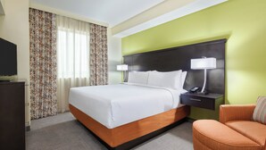 Habitación de hotel con una cama grande, una mesita de noche con lámpara, una silla y un televisor sobre un mueble.
