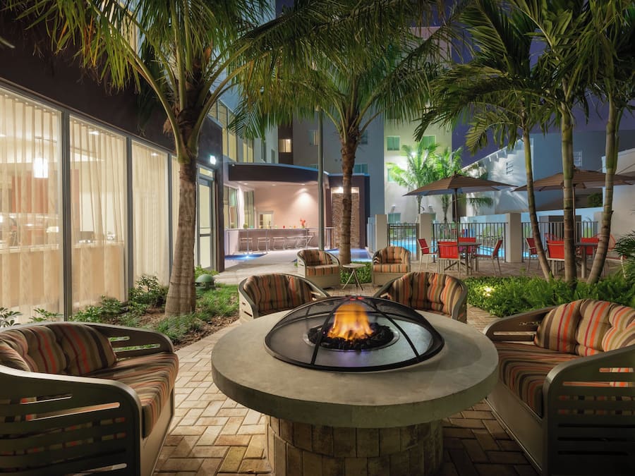 Un área de estar al aire libre con una chimenea, rodeada de palmeras y muebles de exterior modernos.