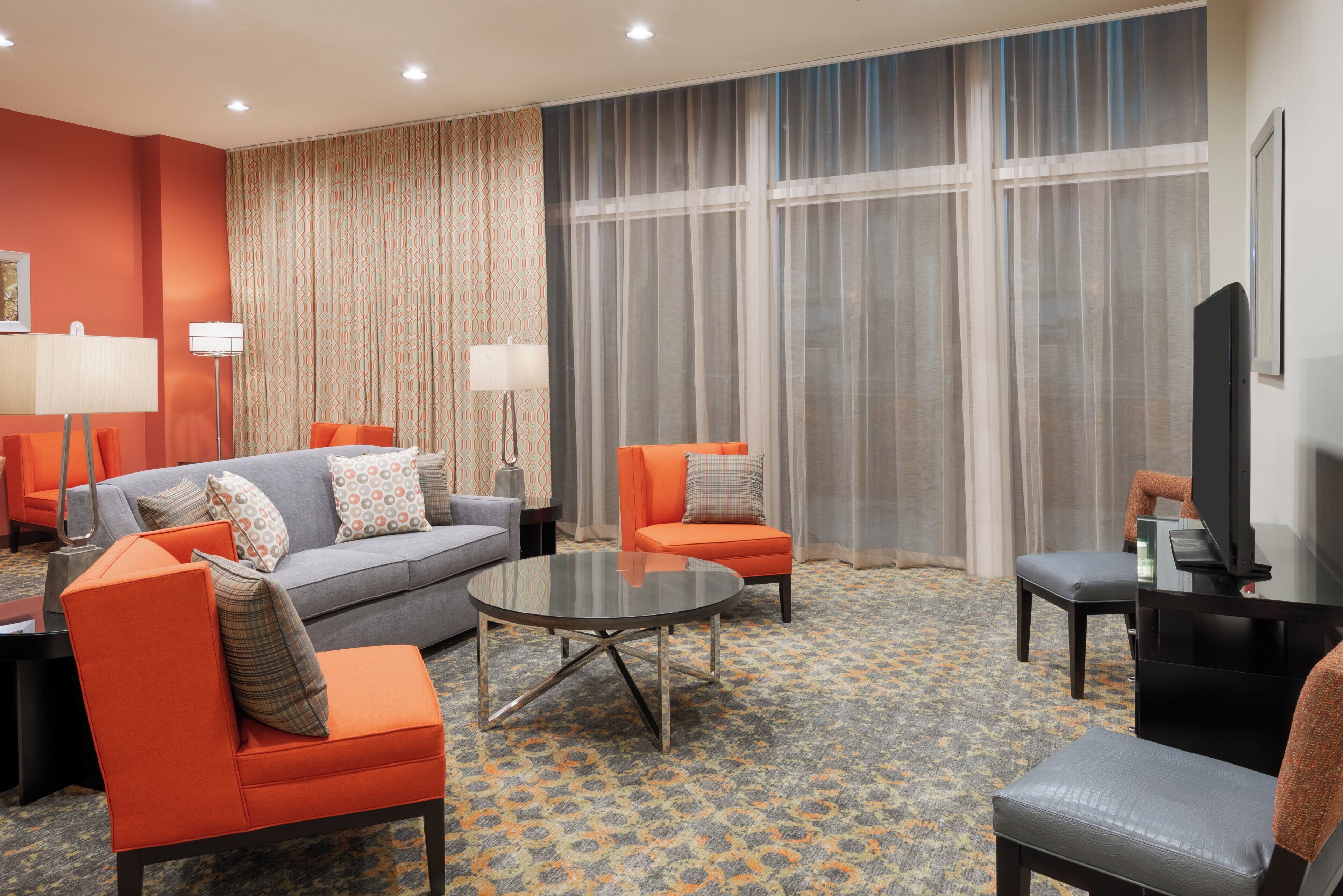 A hotel room with a grey sofa, orange armchairs, a round coffee table, and a television.