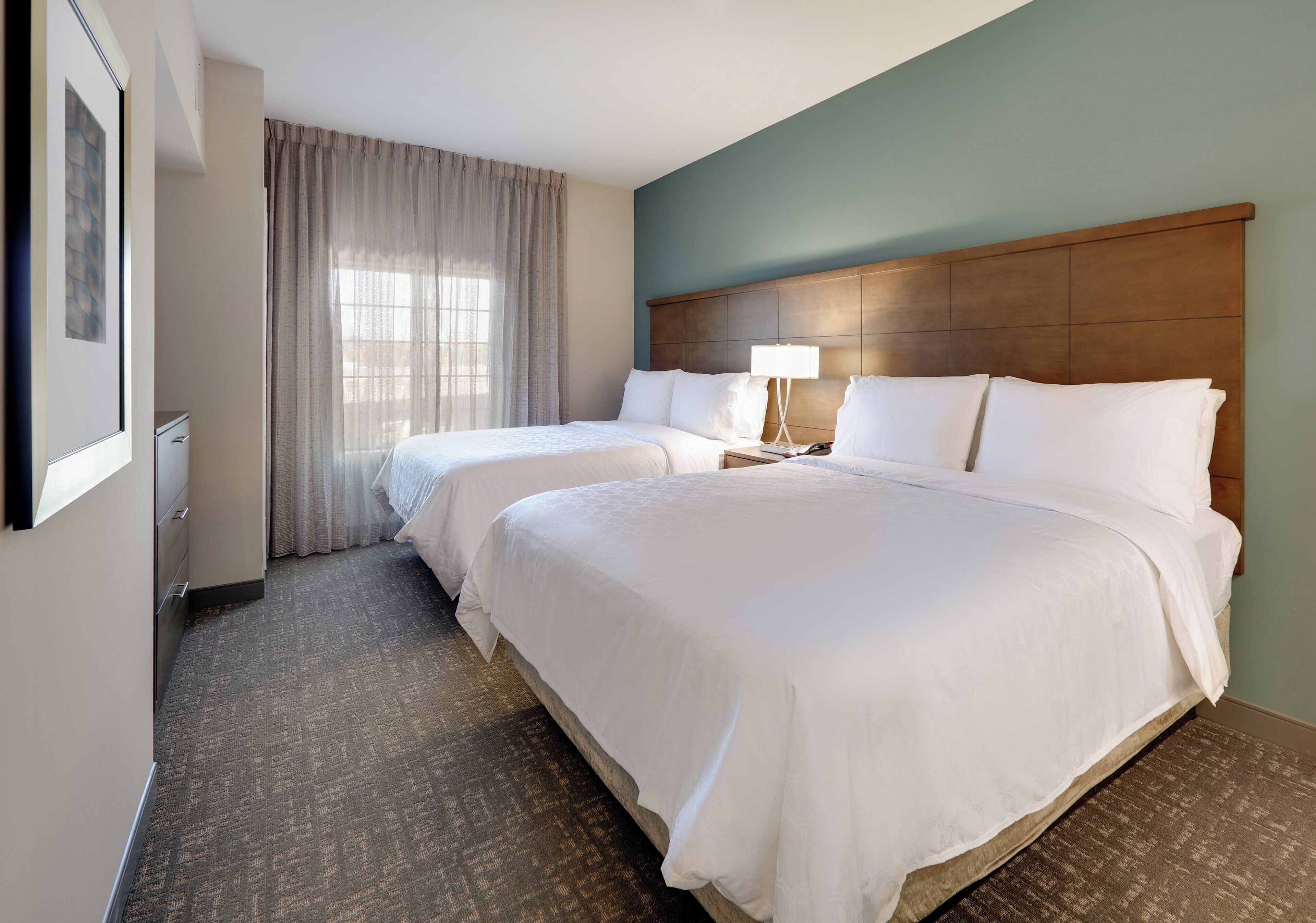 A hotel room with two beds, a wooden headboard, a nightstand with a lamp, a window with curtains, and a framed picture on the wall.