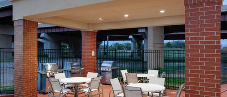 An outdoor patio with brick pillars, metal grill stations, and white tables and chairs.