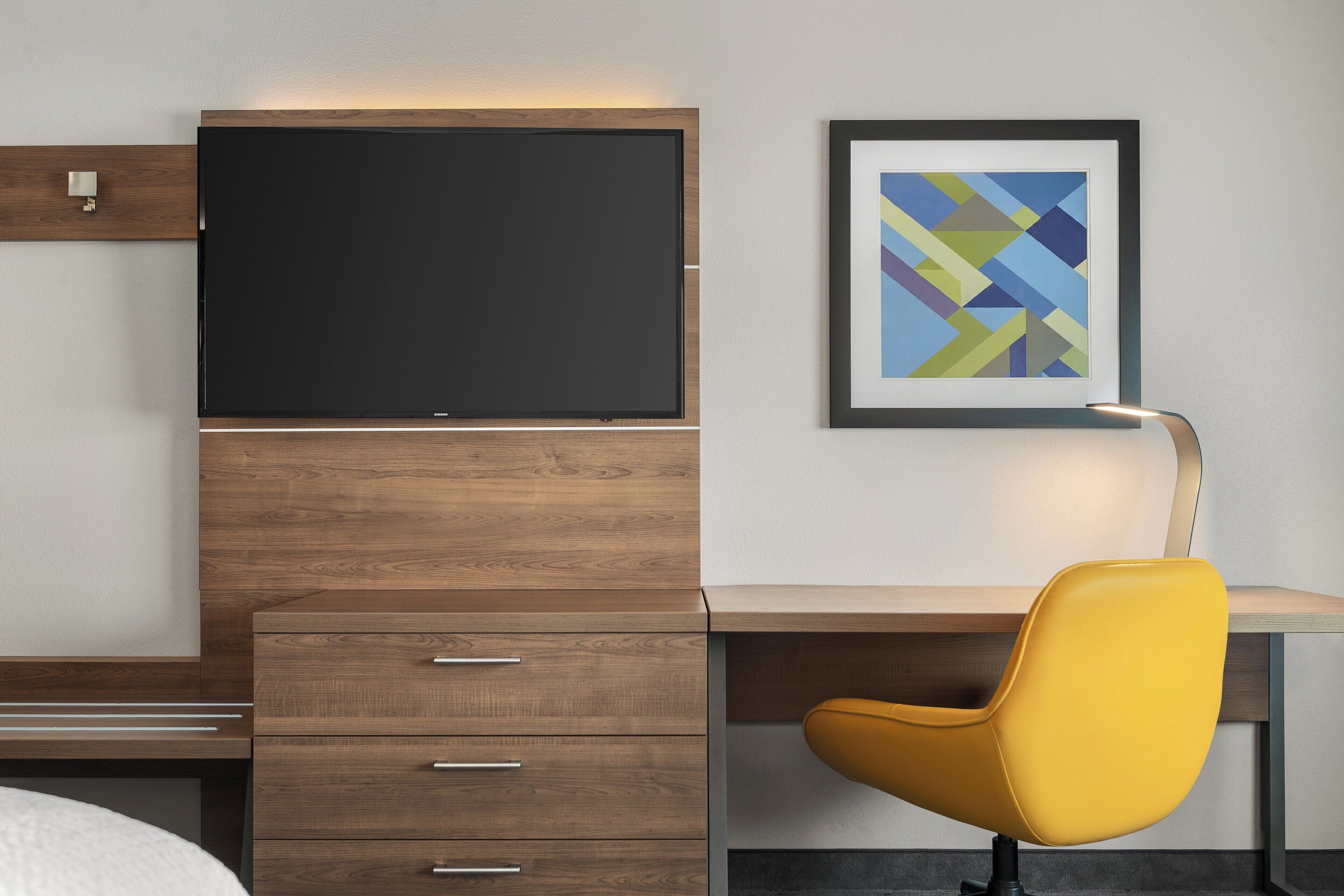 A modern hotel room with a wooden desk, a yellow chair, a flat-screen TV, and a framed abstract art piece on the wall.