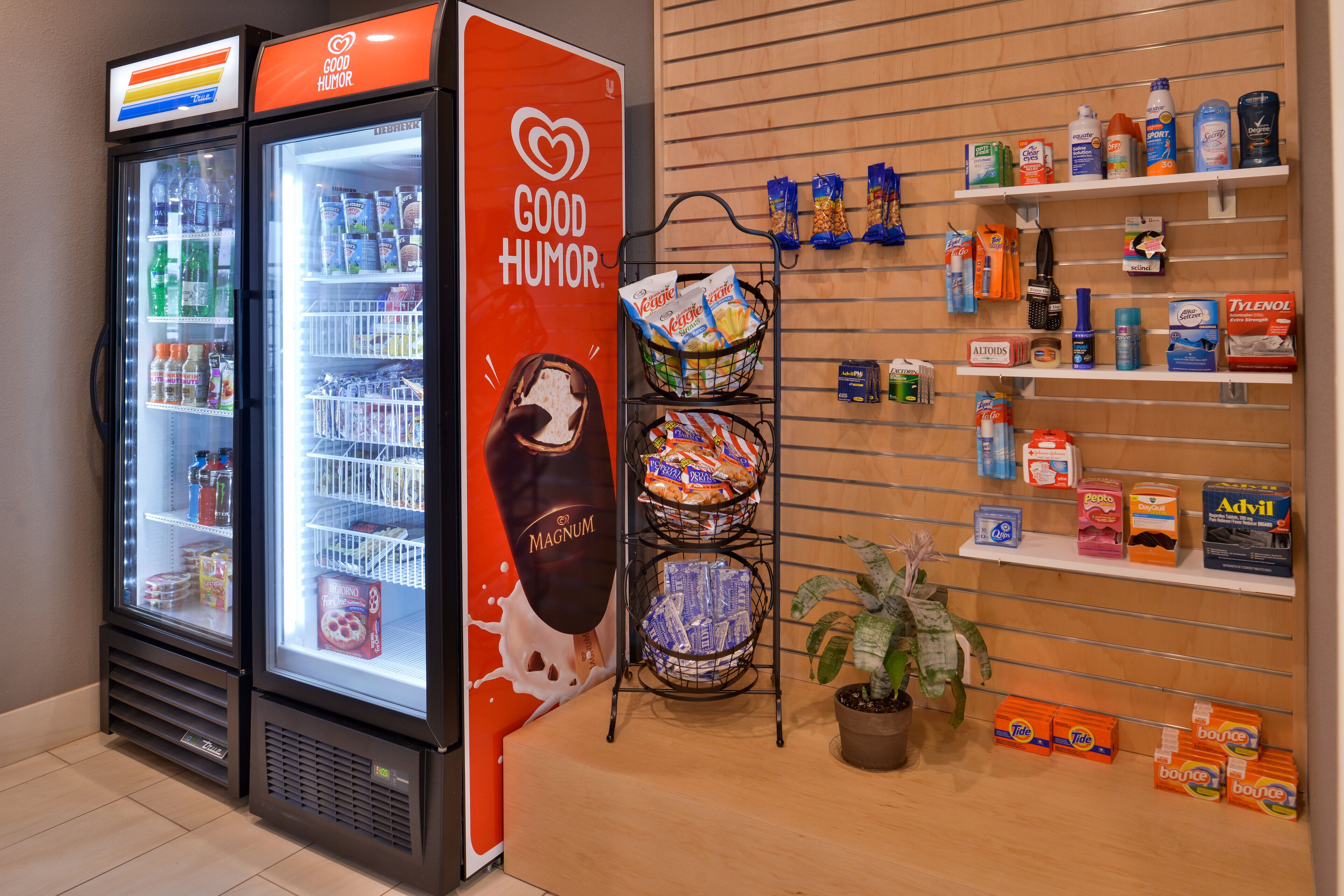 A refrigerated display with various beverages and snacks, a 'Good Humor' advertisement, and shelves stocked with personal care products.
