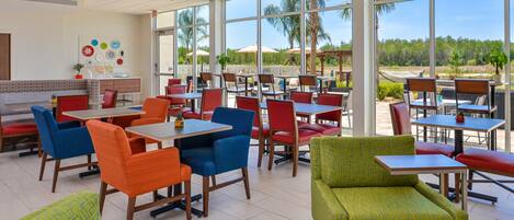 A spacious indoor dining area with multiple tables and chairs, large windows offering a view of palm trees and greenery outside.