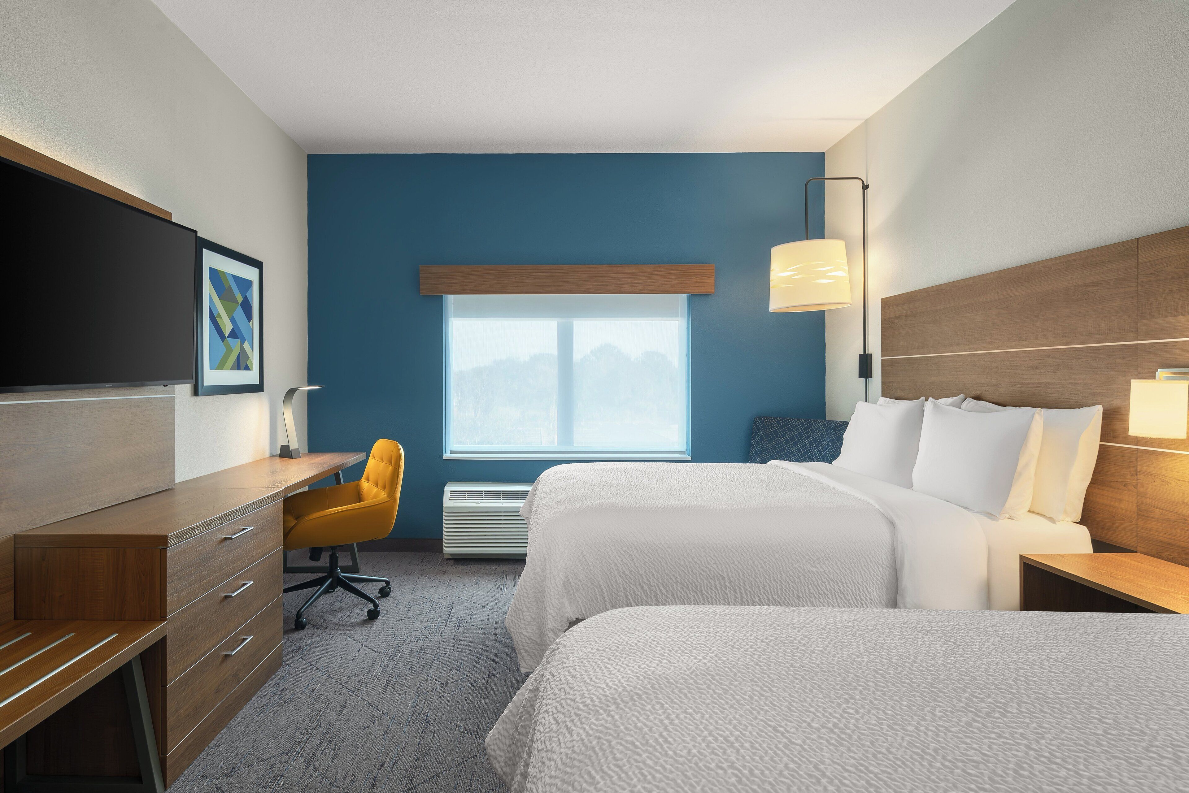 A hotel room with a desk, two beds, a chair, a TV, and a window with a view.