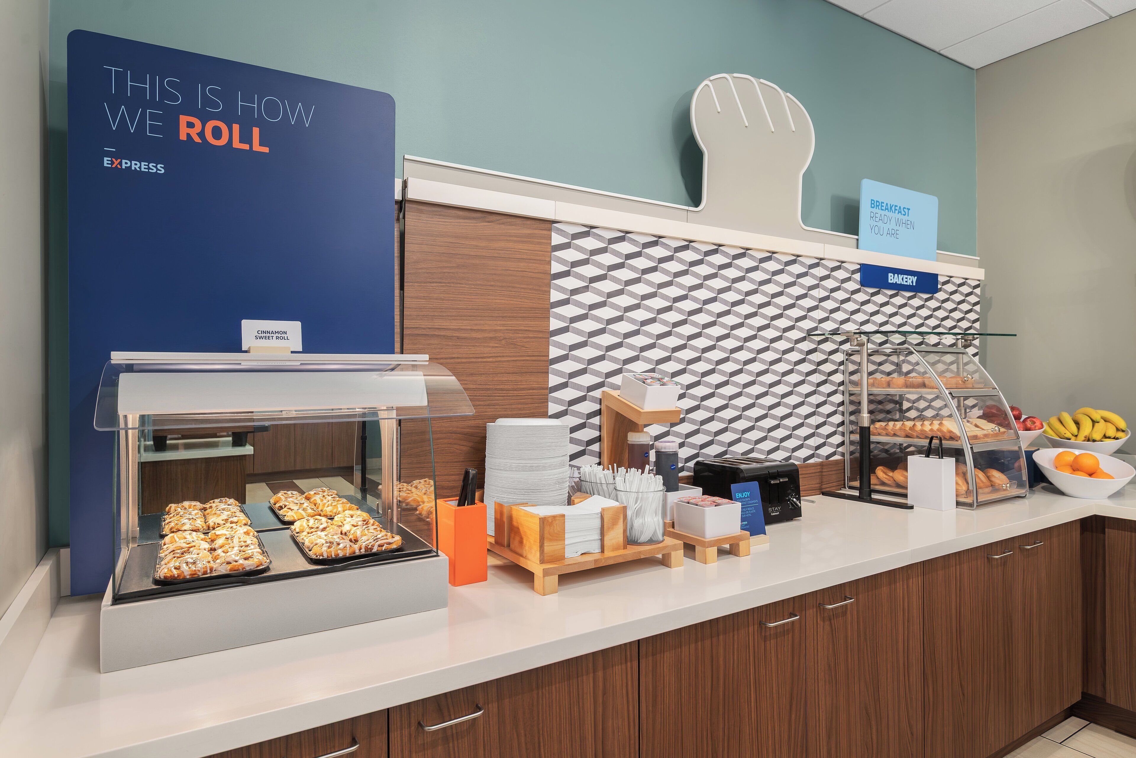 Buffet de café da manhã com uma placa indicando 'THIS IS HOW WE ROLL' e outra placa com a mensagem 'CAFÉ DA MANHÃ PRONTO QUANDO VOCÊ ESTIVER'.
