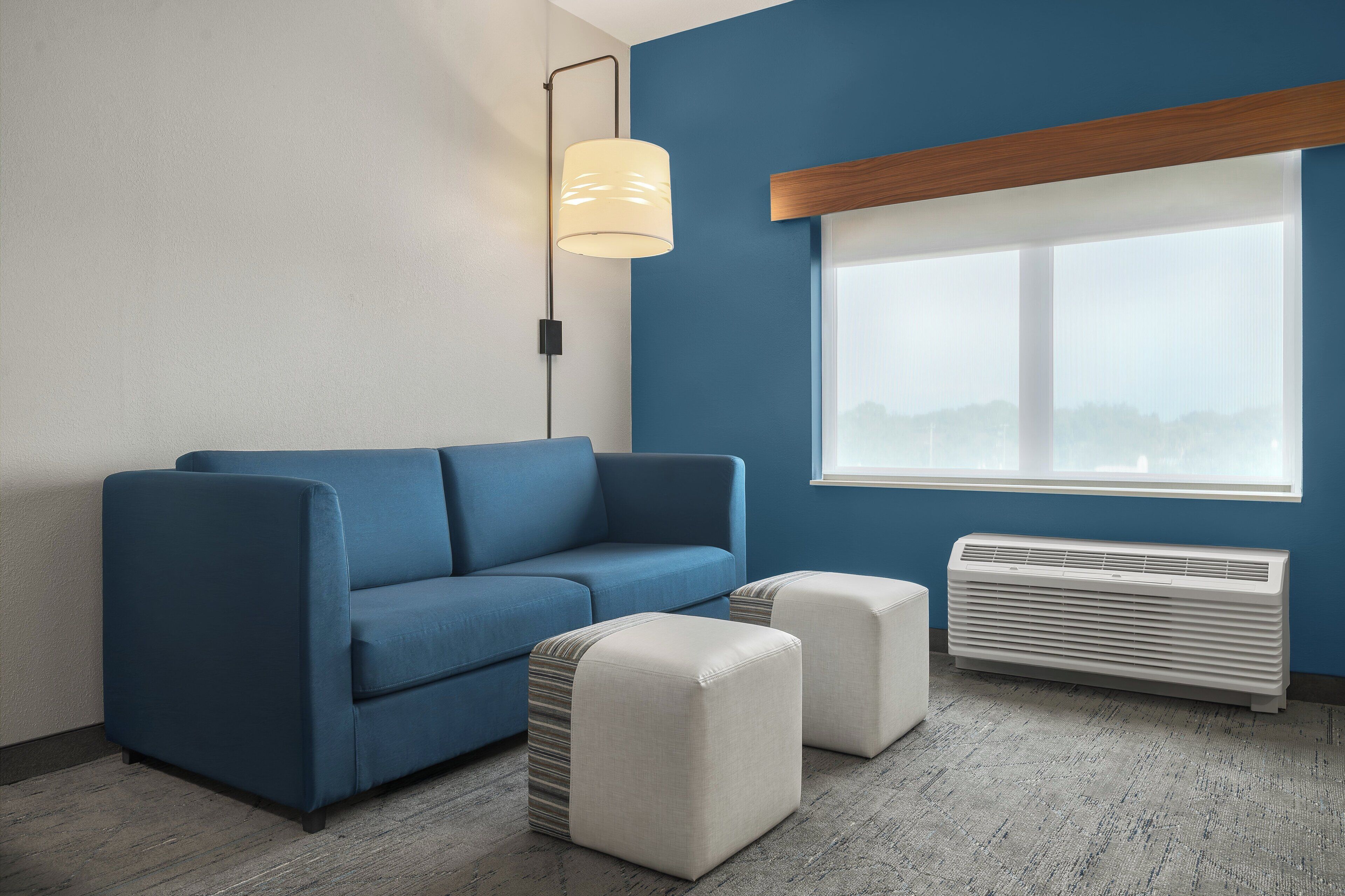 Un salon moderne avec un canapé bleu, deux poufs, une lampe sur pied et une fenêtre avec des stores.