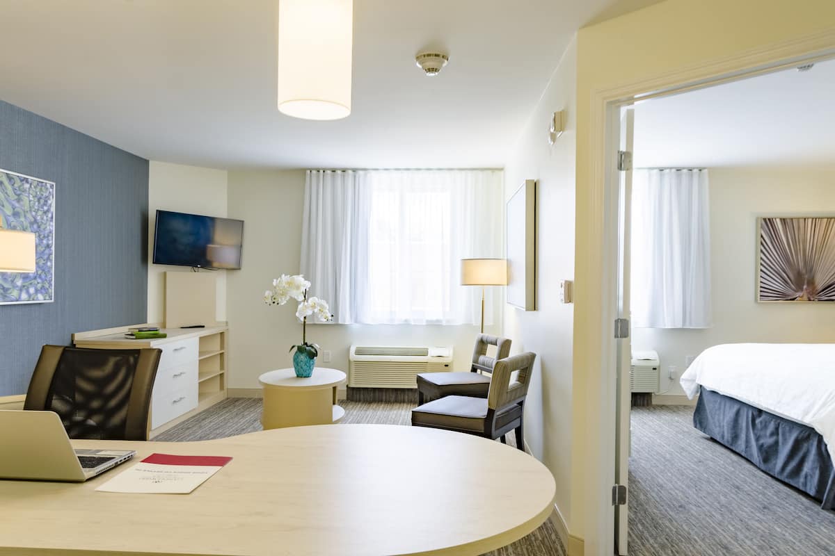 A hotel room with a bed, a desk with a laptop, a chair, a TV, and a window with curtains.