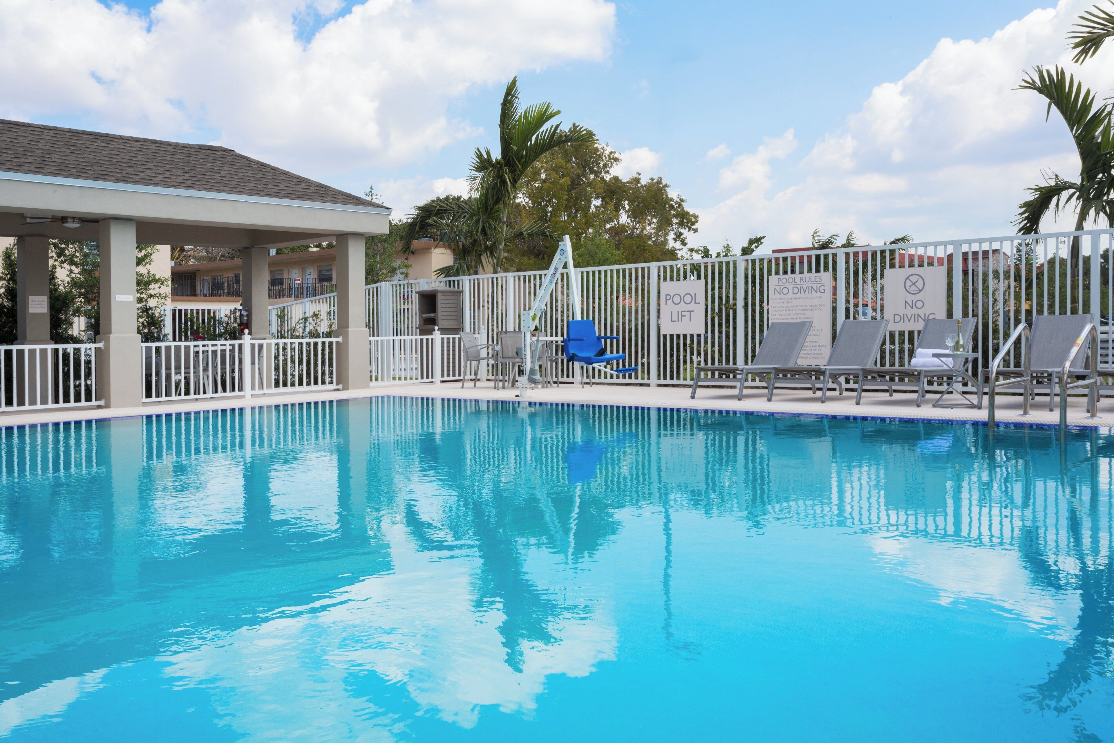 A clear blue pool with a white fence, lounge chairs, and a covered area with a roof.
