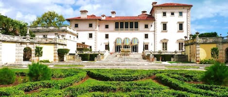 A large, historic building with a symmetrical facade, a central entrance with a staircase, and a formal garden with neatly trimmed hedges in front.