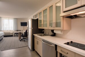 A modern hotel room with a kitchenette, including a stove, sink, and refrigerator. There is a bed, a desk, and a TV in the room.