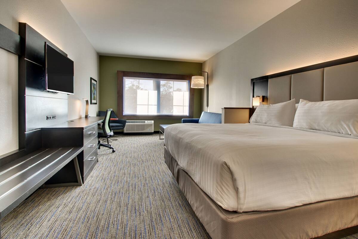 A hotel room with a large bed, a desk with a chair, a flat-screen TV mounted on the wall, and a window with a view of trees.